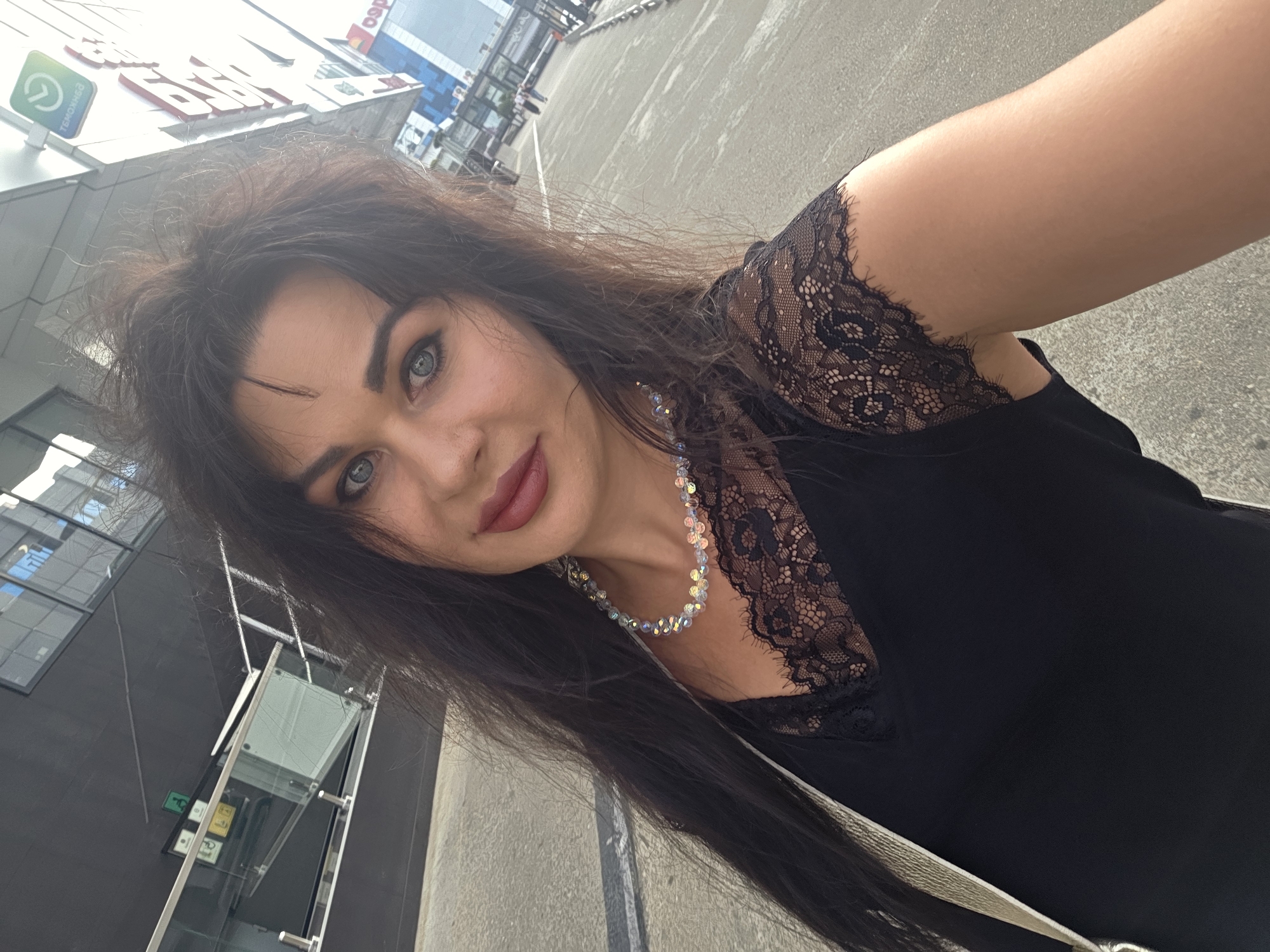 A woman is taking a selfie outdoors in front of a modern building. She has long, dark, wavy hair and blue eyes, with dark makeup on her eyes and lips. She is wearing a black top with lace detailing on the shoulders and neckline, and a beaded necklace. A silver bag is slung over her shoulder. The background shows a concrete walkway and a multi-story building with glass windows and signage. The scene is likely in a commercial or urban area.