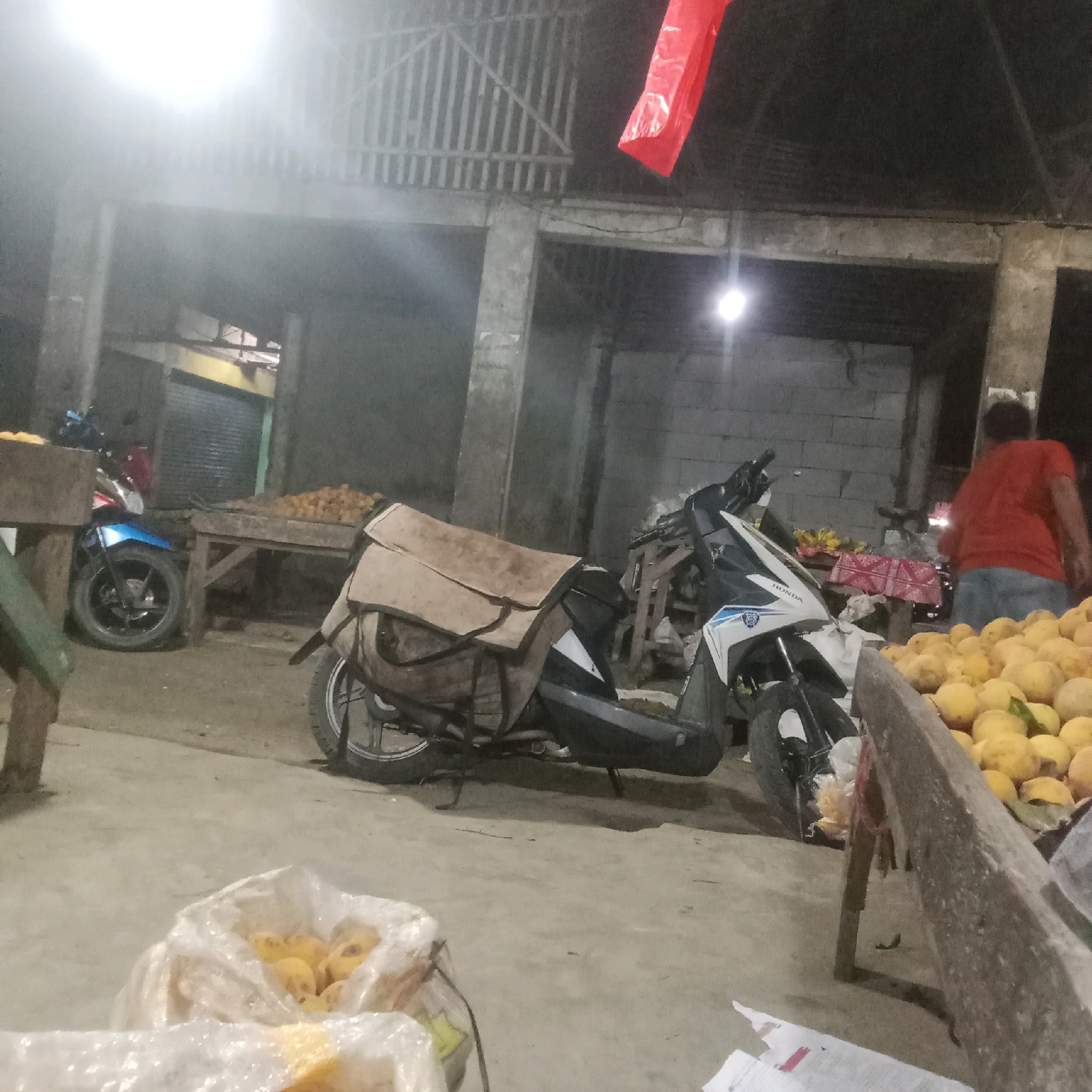 In Karawang, Indonesia, a busy market scene unfolds under the dim illumination of overhead lights. A white and blue Honda motorcycle, equipped with a tan fabric cover over its seat and rear, is parked prominently in the foreground. To the left, another motorcycle, predominantly blue and silver, is partially visible. Several wooden stalls are laden with produce. A large quantity of yellow fruits, possibly mangoes or citrus, are displayed in a wooden crate on the right. Another crate or stall on the left holds a pile of similar yellow fruits. In the background, a person in a red shirt and blue pants stands near a stall adorned with a red and white patterned cloth, and a bunch of bananas. The environment appears to be an open-air market structure with concrete pillars and a corrugated metal roof. The ground is paved with concrete. Two plastic bags filled with yellow fruits are also visible in the lower portion of the image.