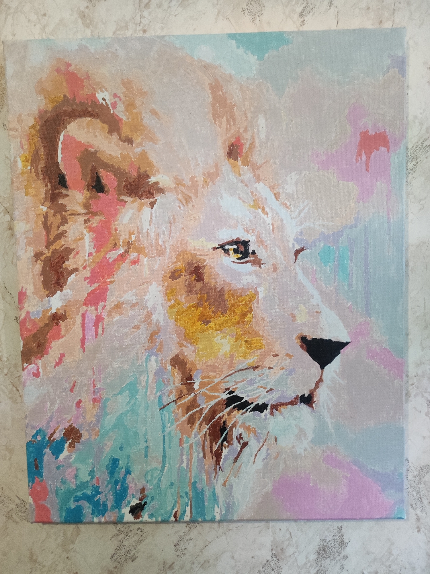 The image shows an abstract painting of a lion's face, rendered in pastel colors with visible brushstrokes. The painting is square and appears to be on canvas. The lion is depicted in profile, facing right, with its mane blending into a background of soft blues, pinks, and creams. The lion's features are loosely defined, with prominent eyes, a black nose, and hints of its mane in shades of brown, pink, and yellow. The background is composed of broad, abstract color blocks and drips. The painting is displayed on a textured surface that resembles marble or textured wallpaper. The image is taken from a slightly overhead angle. The location in the city of Cherkasy, Ukraine, is not discernible from the image itself.