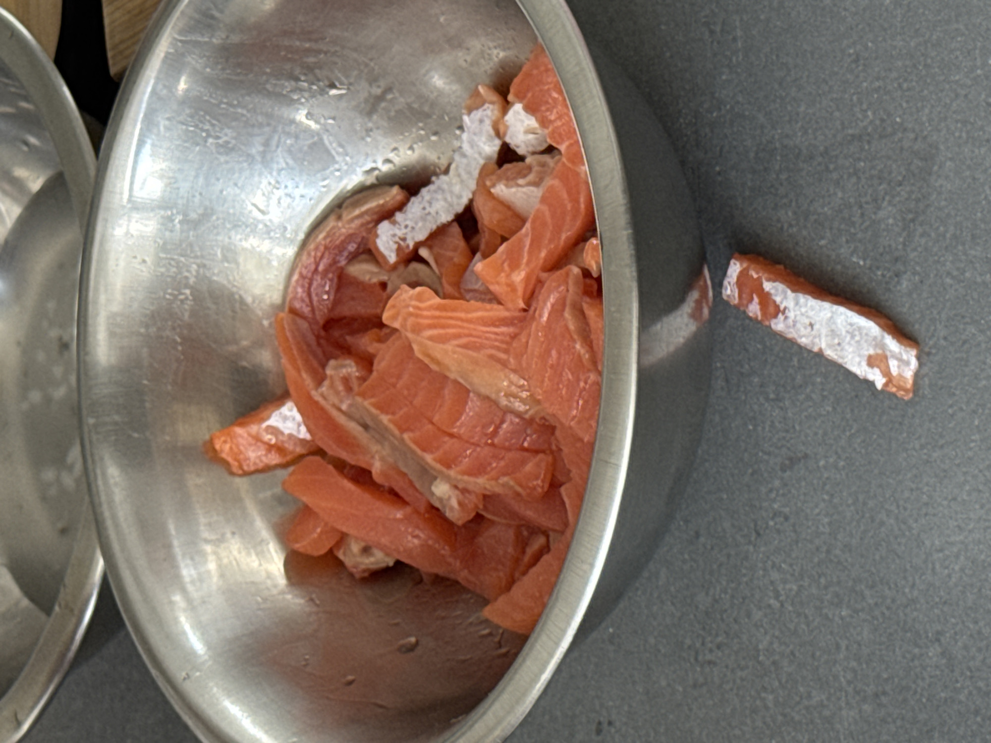 A close-up shot shows a stainless steel bowl filled with raw, sliced salmon. The salmon pieces are a vibrant orange-pink color and appear to be cut into bite-sized strips. Some pieces have white fat running through them. The bowl is slightly wet, with water droplets visible on its inner surface. One piece of salmon has fallen out of the bowl and rests on the dark gray countertop in front of it. The countertop has a speckled texture. In the background, another stainless steel bowl is partially visible, out of focus. The lighting suggests an indoor setting, possibly a kitchen. There are no people visible in the image. The image is taken from a slightly high angle, looking down into the bowl.