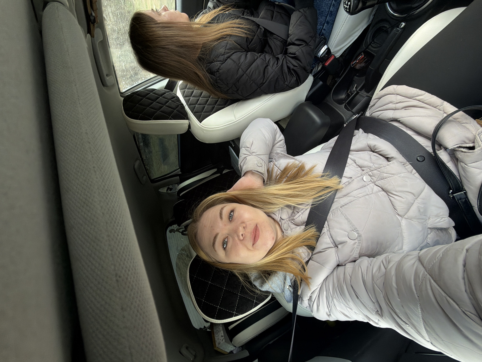 This media file, set in Noviy, Russia, is oriented on its side, depicting the interior of a car with two women.
In the foreground, a blonde woman with blue eyes looks directly at the camera, offering a slight smile. She is wearing a light gray quilted jacket and a seatbelt, and her head rests on a black and white quilted headrest. A black shoulder bag strap is visible on her right.
In the background, another woman with long brown hair, wearing a black quilted jacket and seatbelt, is seated. Her face is turned towards a car window, looking out. The window appears to be wet, suggesting rain or recent precipitation outside.
The car's interior features black and white quilted seats and headrests, contrasting with the plain gray fabric of the seat in the far left. A central console with a gear stick and other car controls is visible between the two front seats. A faint "PILOT" text is visible on the side of one of the seat supports near the blonde woman. The overall impression is of a casual journey in cool, possibly rainy, weather.
