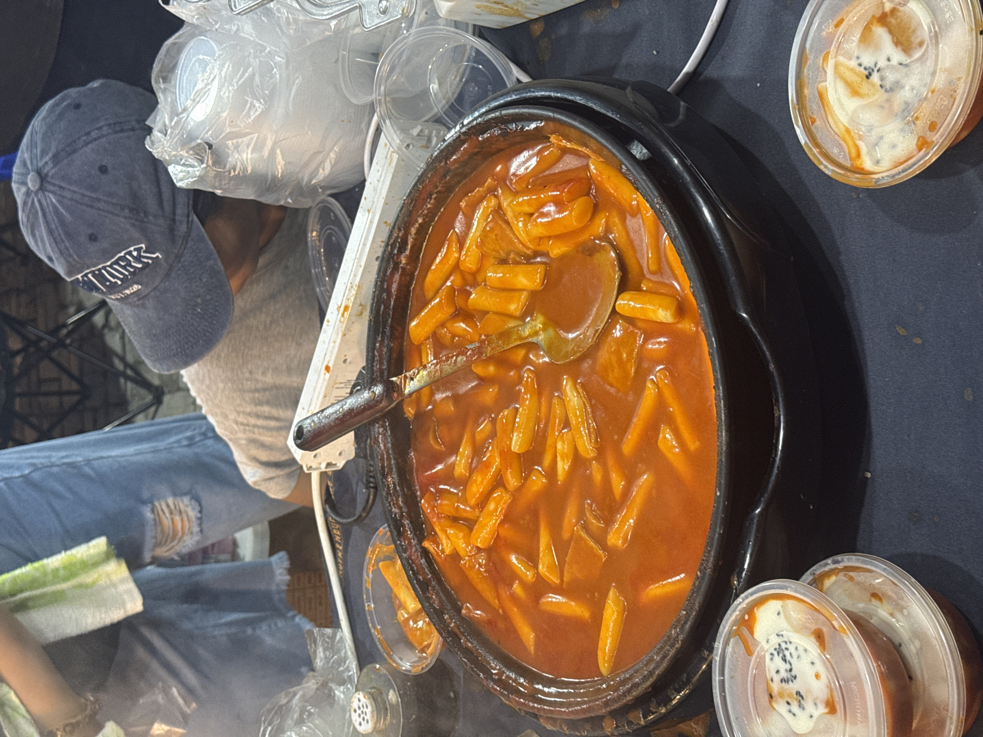 The image depicts a close-up, eye-level view of a large pot of Tteokbokki, a popular Korean street food, at an outdoor food stall. The Tteokbokki, which consists of chewy rice cakes in a vibrant red, spicy sauce, is being served with a ladle. Several clear plastic containers with lids, some containing smaller portions of the dish or dipping sauces, are visible in the foreground. In the background, a person wearing a grey t-shirt and a blue baseball cap with "NEW YORK" written on it is leaning over the stall, partially obscured by a plastic bag. Another person is visible to the left, wearing ripped denim jeans and holding a green and white striped cloth. The environment appears to be a bustling market or street food setting, possibly at night, given the lighting and the presence of other indistinct stalls or tents in the background. The overall scene suggests a casual, informal food vending operation. This scene is typical of food stalls found in popular night markets and street food areas of Davao City, Philippines, where diverse international cuisines are readily available.