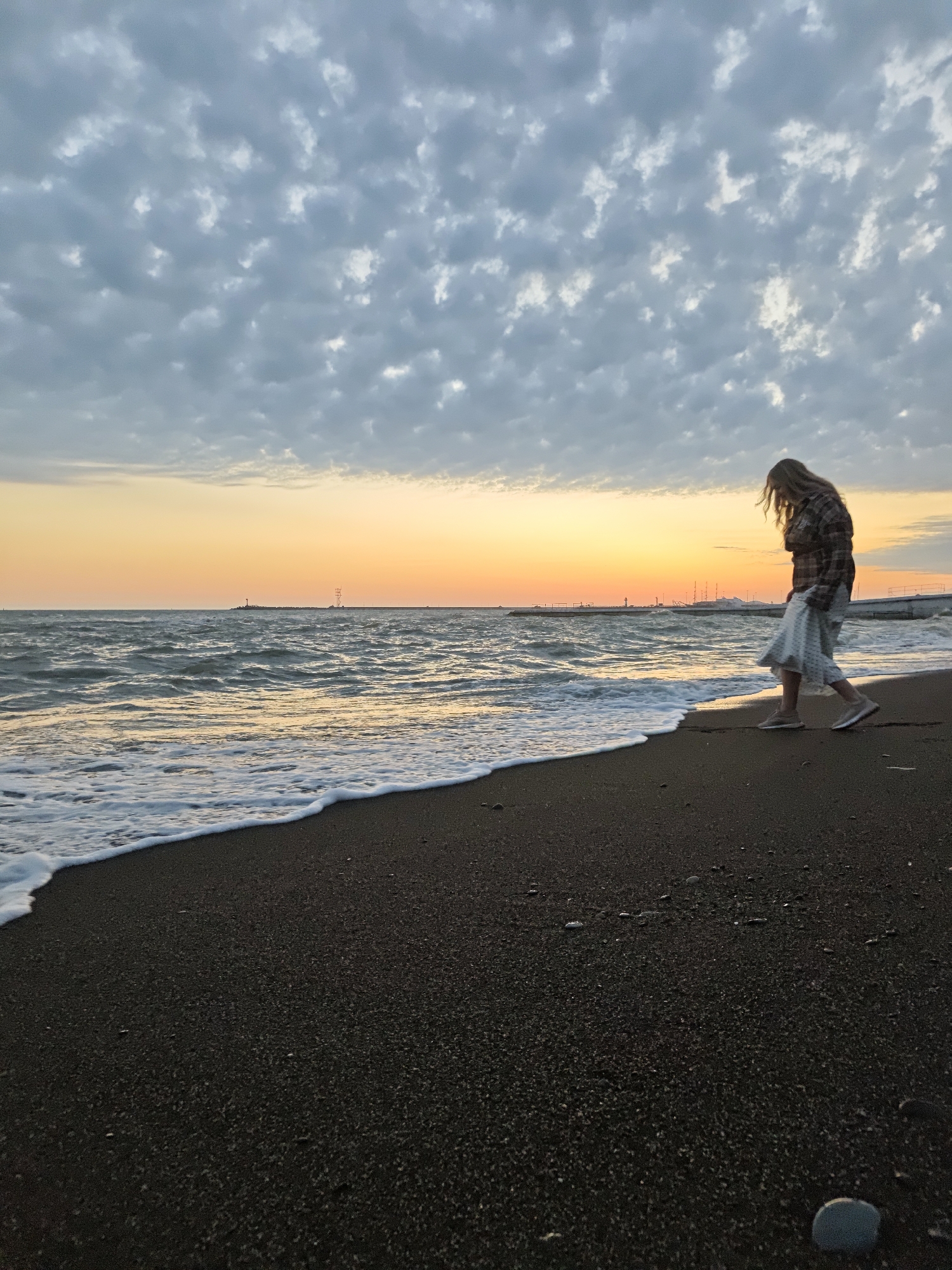 A woman is walking on a beach in Sochi, Russia. The sky is cloudy with an orange glow from the sunset. The sea is wavy and has white foam where it meets the black sand beach. The woman is wearing a plaid shirt and a light-colored skirt. She is walking towards the right of the frame and appears to be looking down. In the distance, there are structures in the water, possibly a breakwater or pier. A small rock sits near the bottom right corner of the frame.