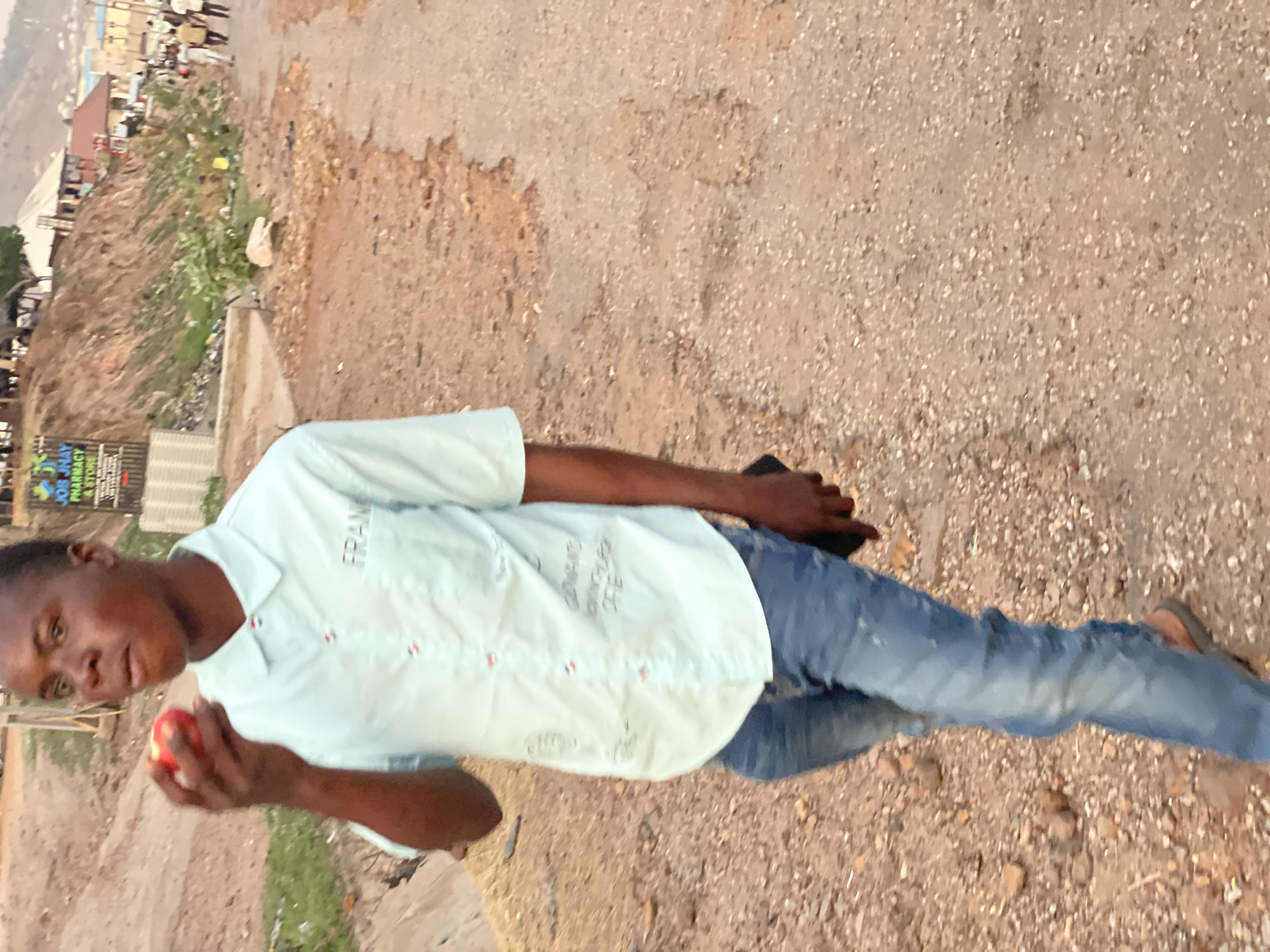The image shows a man outdoors in Lokoja, Nigeria. He is holding a red object, likely an apple. He is wearing a light blue button-down shirt, jeans, and sandals. He appears to be standing on a dirt or gravel road. In the background, there is a hillside with some buildings and signs. The environment suggests a location on the outskirts of Lokoja.