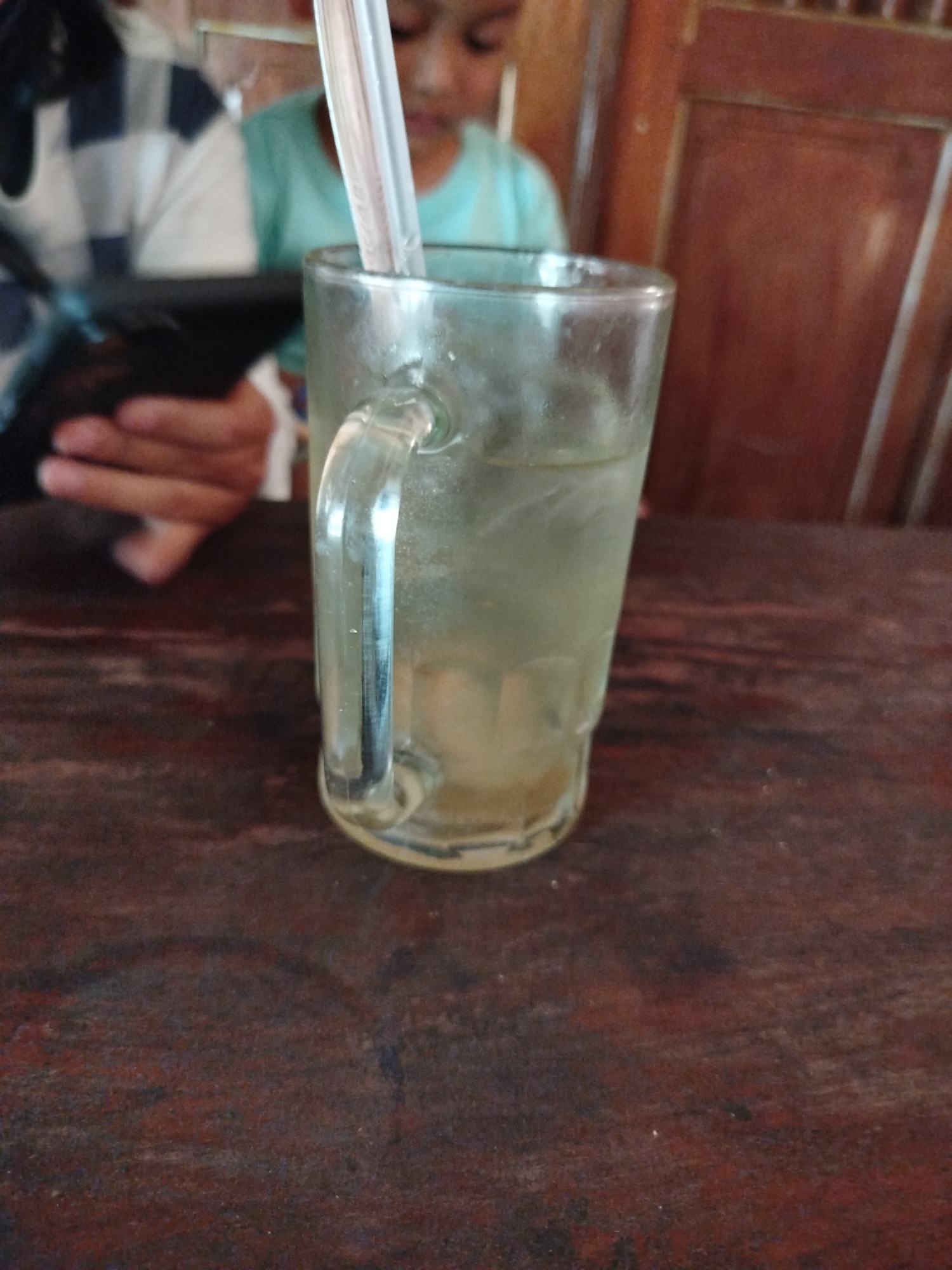 A glass of iced water with a straw sits on a dark wooden table in Pojok, Indonesia. The glass has a clear handle and is filled with water and ice. Behind the glass, a person wearing a blue and white checkered shirt is holding a black object, possibly a phone. To the right of that person, another individual in a turquoise shirt is visible. The background consists of a wooden wall and chair. The scene appears to be a casual gathering or meal setting.