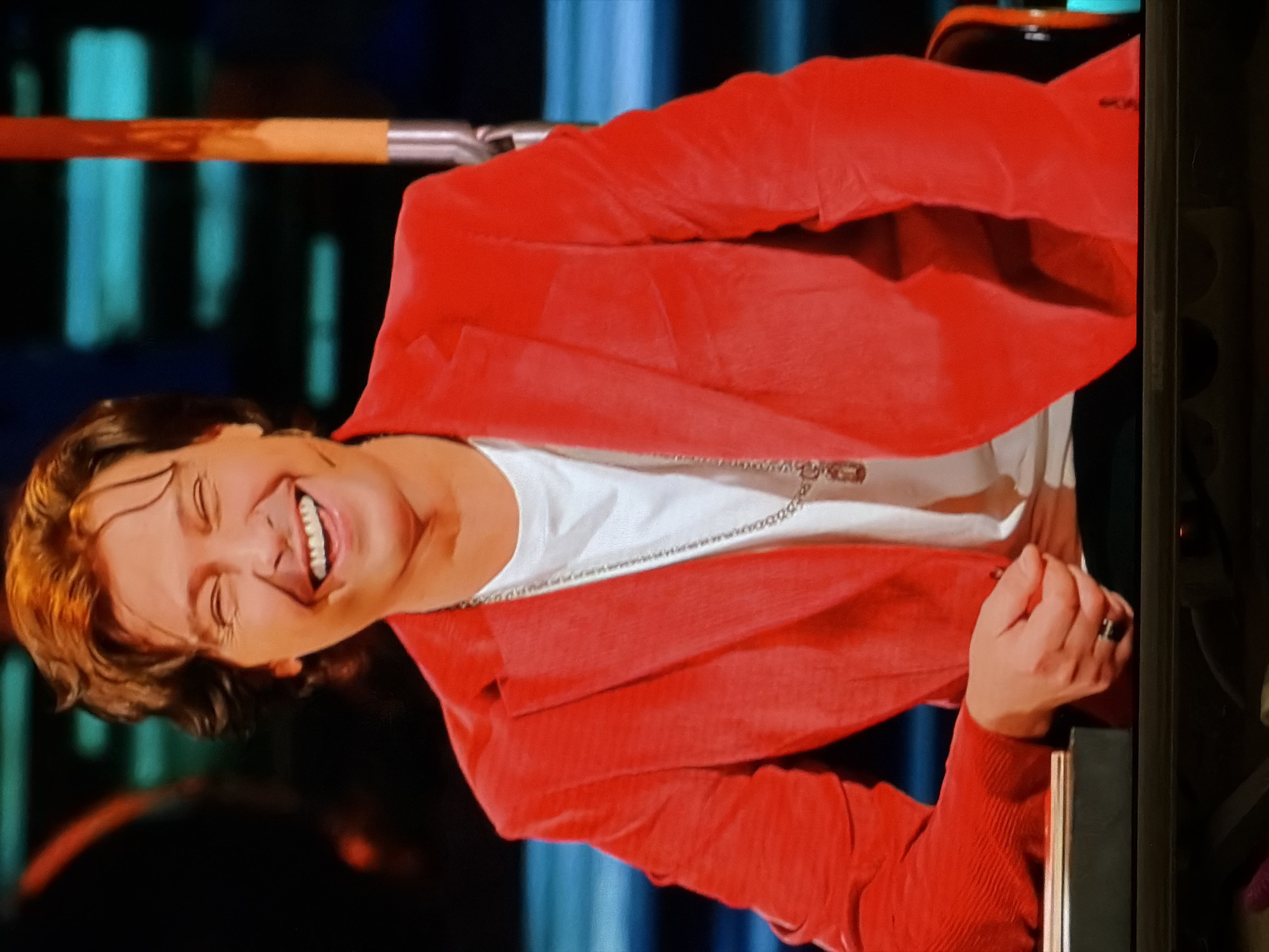 A man is smiling, wearing a red jacket and a white t-shirt with a chain necklace. He is in an indoor setting with dark backgrounds. A piece of a wooden or metallic object is visible in the upper left corner. The image does not contain enough information to confirm the location as Moscow, Russia.