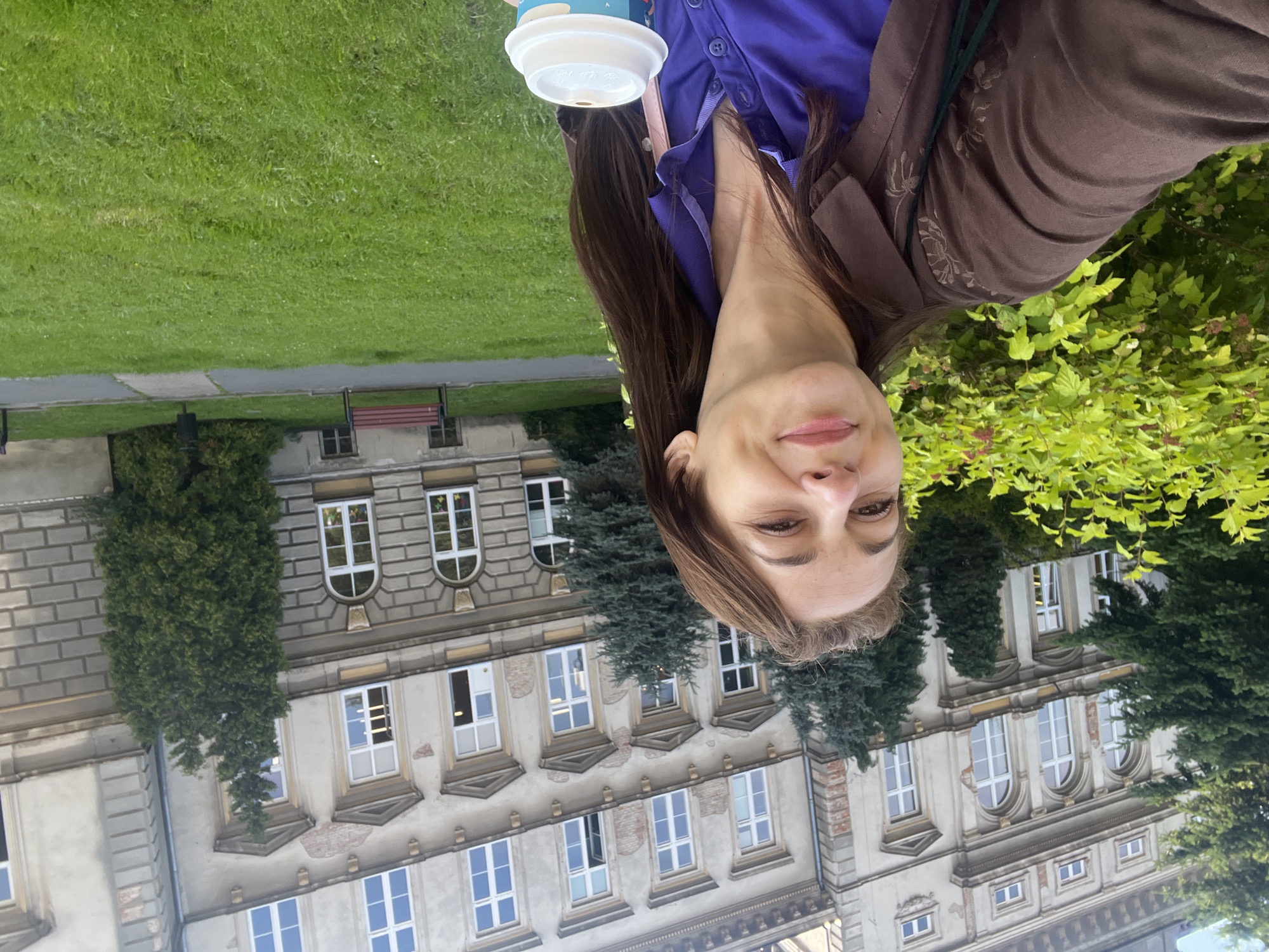 The image shows a young woman holding a disposable cup. She is wearing a purple shirt and a brown jacket. Her hair is long and brown. She is smiling at the camera. The background is out of focus, but it appears to be an outdoor setting with a building and greenery. The image is taken from a low angle, and the woman is positioned in the upper half of the frame. The lighting suggests it is daytime. There is no visible text in the image. The woman's expression appears happy and relaxed. The building in the background seems to be of historical or academic architecture, with multiple windows and classical detailing. Lush green grass and dense foliage of trees are also visible.
