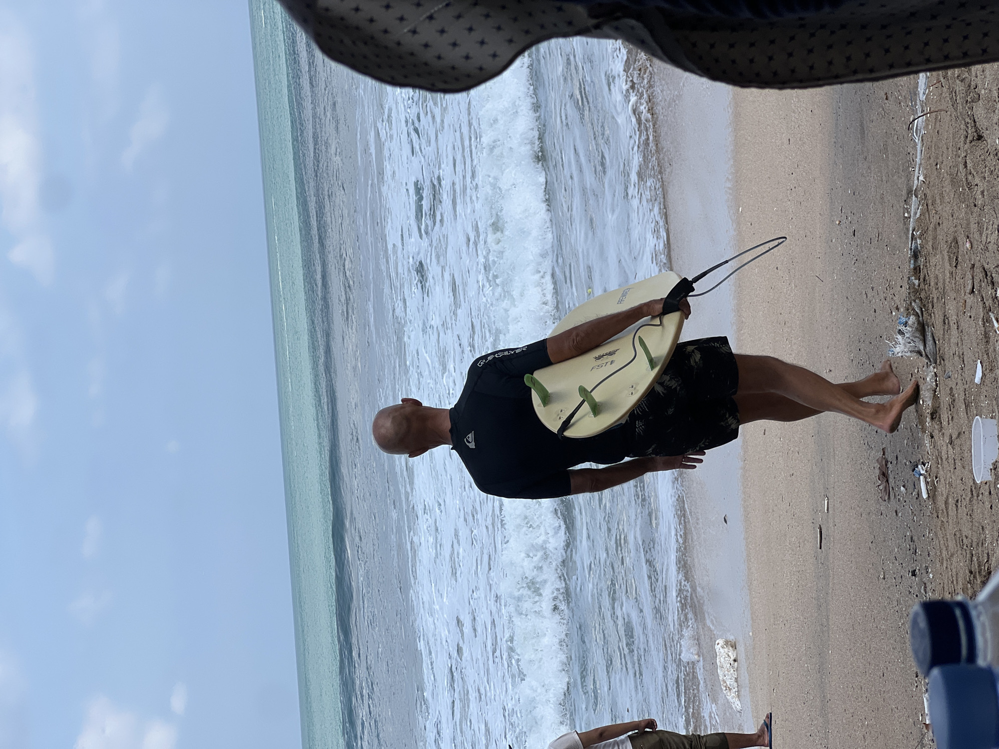 A bald man wearing a black Quiksilver wetsuit and black patterned shorts walks on a sandy beach in Kuta, Indonesia, carrying a pale yellow surfboard with green fins. He is walking away from the viewer towards the ocean, which has moderate waves and white foam. The beach is littered with debris, including plastic bottles and other trash. In the background, the ocean meets a cloudy sky. To the left, another person is partially visible, wearing light-colored shorts and flip-flops. To the right, a person is mostly obscured by the foreground, wearing a dark garment with a patterned design. The overall scene depicts a beach environment with a surfer preparing to enter the water, amidst a somewhat littered shoreline.