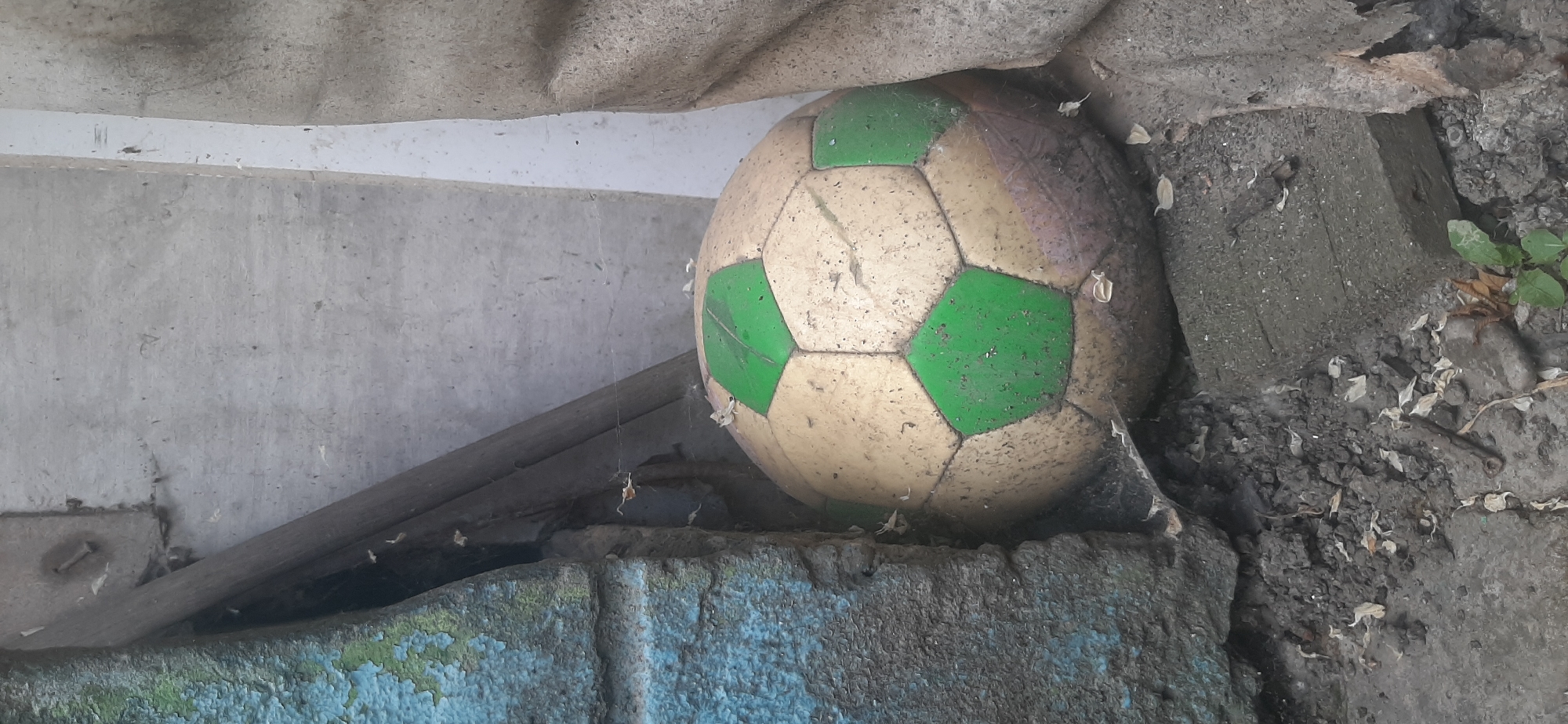 The image shows a dirty, weathered soccer ball with green and white panels resting on a rough, weathered concrete surface. The ball is positioned at an angle, partially obscured by other debris and the surrounding environment. Cobwebs are visible around the ball and hanging from above, suggesting it has been in this location for some time and is perhaps neglected.
The scene appears to be outdoors in a somewhat dilapidated or neglected area. The background consists of a rough, grey concrete wall and a rough, textured surface that could be a roof or overhang. There are also loose pieces of debris, including dried leaves and dirt, scattered around the ball and on the ground. The lighting suggests it is daytime, but the overall atmosphere is one of disuse and abandonment. There are no people visible in the image. The location is identified as Yessentukskaya, Russia. No text is visible in the image.