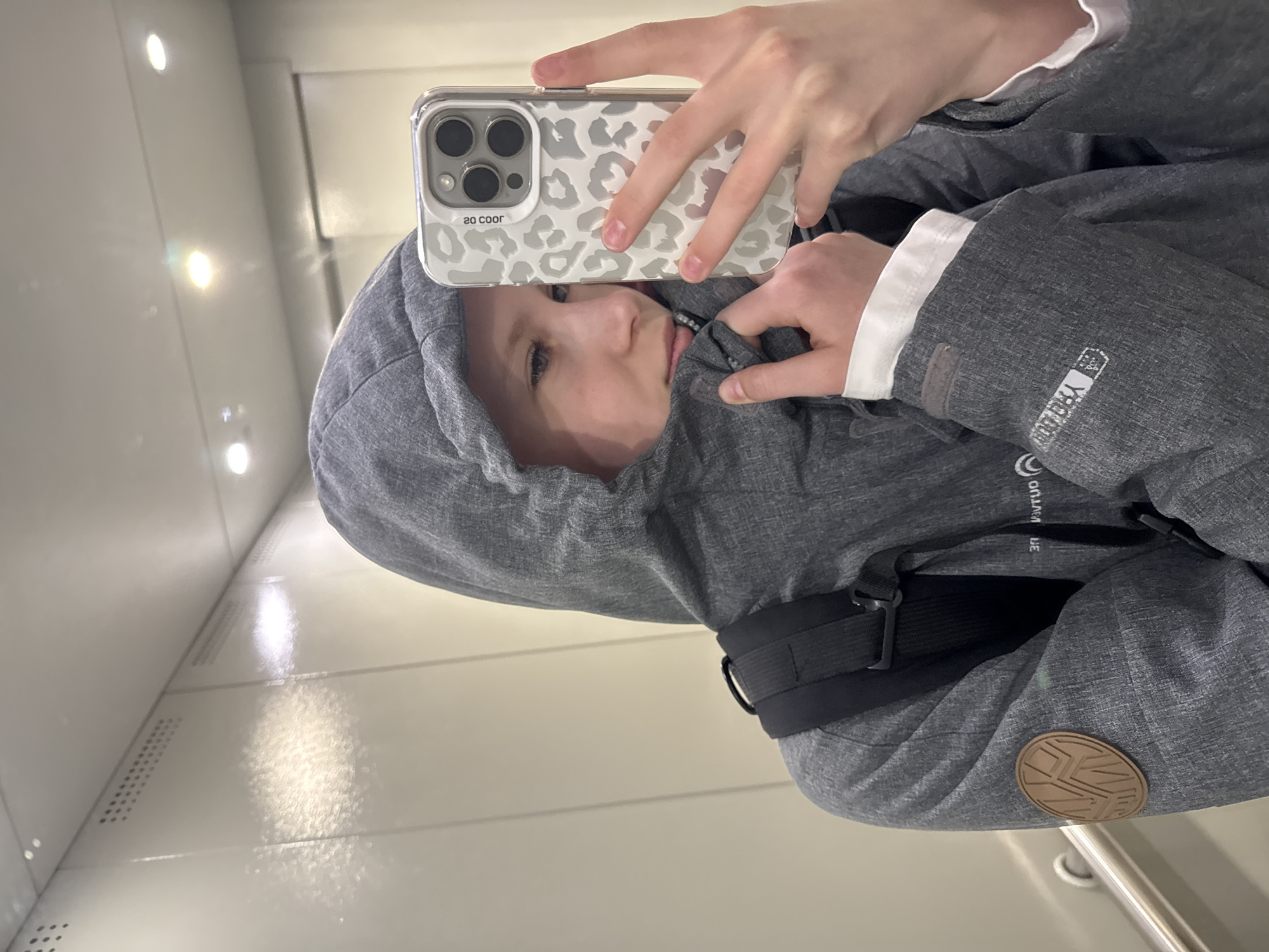 A person is taking a selfie in what appears to be an elevator. The individual is wearing a gray hooded jacket with a black strap across their chest. They are holding a phone with a leopard-print case. The interior of the elevator is metallic and reflective. There is no information in the image indicating that it was taken in Chaikovsky, Russia.