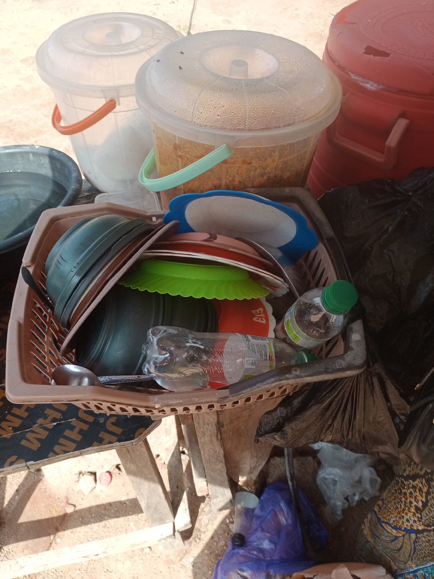 The image depicts an outdoor scene, likely in Jalingo, Nigeria, showing a collection of items arranged on and around a makeshift wooden stand. In the foreground, a brown plastic basket overflows with various kitchenware, including dark green and brown bowls, colorful plates, and a ladle. A partially empty plastic bottle with a green cap is also situated within the basket, alongside other items that appear to be food containers or packaging. To the left, a dark grey basin holds water. Behind these items, several large plastic buckets are visible. One is white with an orange handle, another is translucent with a green handle and contains what appears to be food, and a third is red. The ground beneath the stand is covered in sand and debris, with some scattered objects including a red bottle cap. The overall impression is one of domestic or commercial storage in an informal setting, potentially related to food preparation or sale, given the presence of food containers and the outdoor location. The image is taken from a slightly elevated, angled perspective, with soft lighting suggesting daytime.