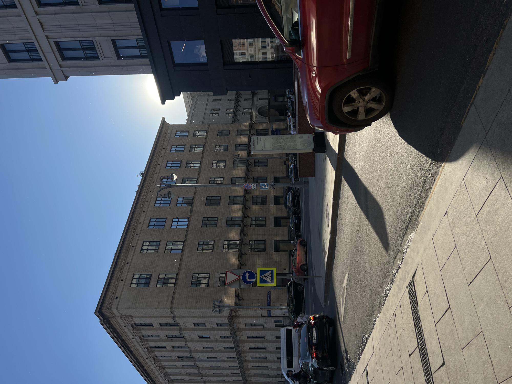 The image captures an urban street scene on a bright, sunny day in Moscow, Russia, specifically at or near the intersection of 1-y Tverskoy-Yamskoy Pereulok (1st Tverskoy-Yamskoy Lane). The sun is positioned low and to the right, causing significant lens flare and casting long, distinct shadows across the asphalt road and sidewalk. Numerous small, bright particulate specks, consistent with dust on a camera lens or atmospheric dust illuminated by strong backlight, are visible in the sky.
On the left side of the frame, a large, multi-story building constructed of light-colored stone or stucco dominates the background. This building exhibits classical architectural elements, including numerous rectangular windows with dark frames, pilasters, and cornices. It is identifiable as the building housing the Russian Ministry of Economic Development.
To the right, a darker, possibly black, modern commercial building facade is partially visible in the foreground, with large reflective windows. Behind it stands a taller, lighter-colored modern building featuring vertical architectural fluting and multiple windows. The intense sunlight creates a strong glare point above the dark building.
The foreground features a light-colored paved sidewalk with rectangular tiles and a metal drainage grate. Adjacent to the sidewalk is an asphalt road, which appears damp or reflective in patches near the curb. A white arrow indicating a right turn is painted on the road surface. A red Jeep Grand Cherokee is partially visible on the right, facing away from the viewer, with its front right wheel and part of its side in the frame. Its dark shadow extends into the road.
On the left side of the road, a black Toyota Camry sedan (license plate T343BA 797, Moscow region code 797) is stopped or moving slowly. Behind it, a white van is partially visible. Two pedestrians are positioned near the black Camry; one appears to be walking, while the other is near the vehicle's rear door. Further down the street, a red sedan is visible.
Several street signs are mounted on a pole near the center of the frame: a red triangular "Give Way" sign, a blue circular "Mandatory Right Turn" sign, and a blue square "Pedestrian Crossing" sign. A multi-headed street lamp stands to the left of these signs. An informational pillar with Cyrillic text and a map-like graphic is situated on the sidewalk near the red SUV. In the far background, additional buildings and a group of people are discernible.