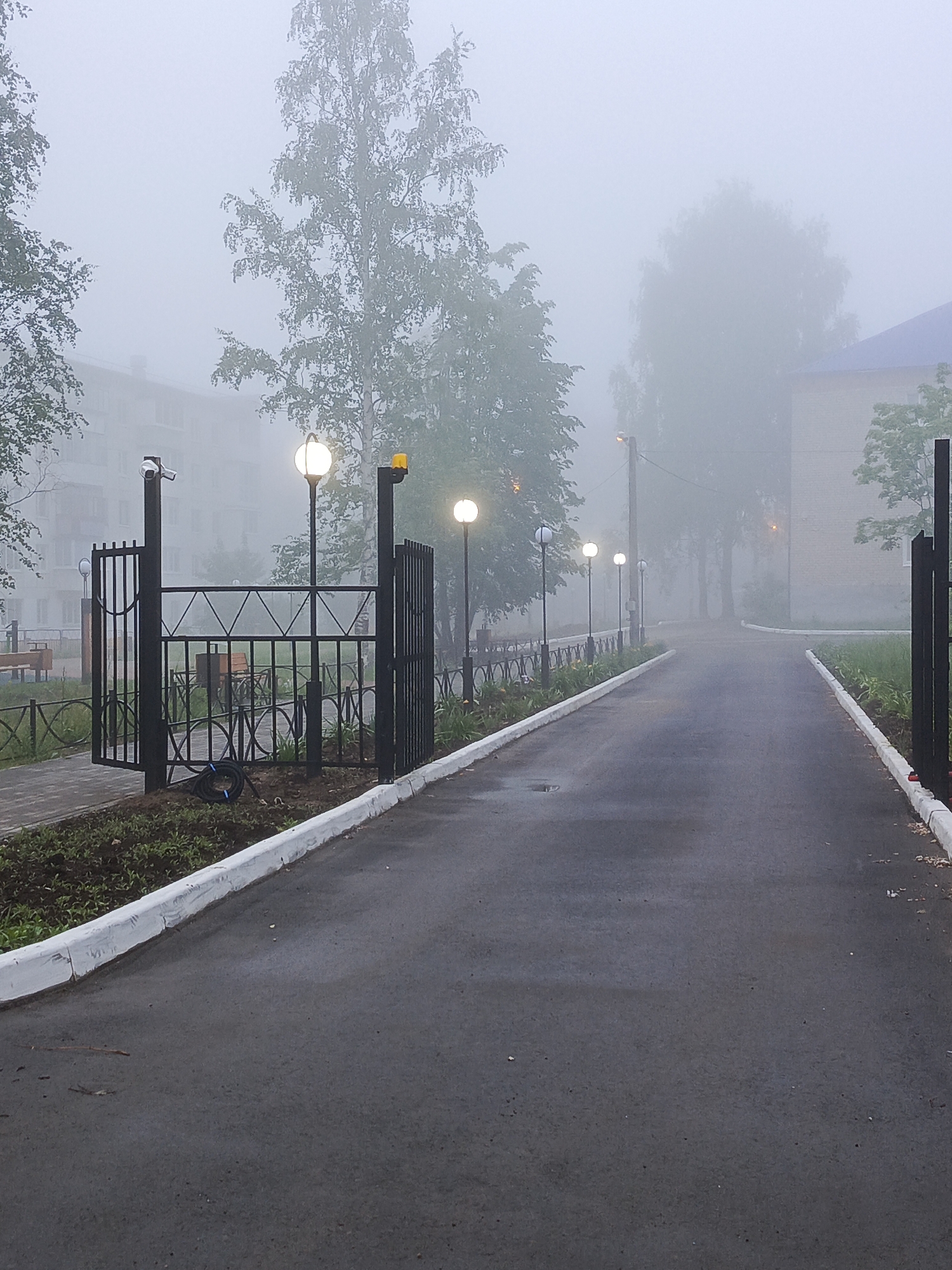 The image depicts a quiet, foggy street or pathway in Chaikovsky, Russia, likely during an early morning or late evening due to the pervasive mist and illuminated streetlights. A paved asphalt road, appearing damp, stretches into the distance, bordered by crisp white curbs.
On the left side, a black metal gate structure stands open, featuring a security camera mounted on its top left post. Beside it, a streetlight with a glowing spherical lamp illuminates the scene, accompanied by a yellow warning light on a taller pole. Beyond this gate, the outlines of buildings and trees are heavily obscured by the dense fog, but a park bench is faintly visible. A coiled black cable lies on the ground near the gate base.
The road itself is lined with a series of similar streetlights on the right side, their spherical lamps emitting a warm glow that cuts through the thick mist. Further along the road and to the right, another black metal gate structure is partially visible in the foreground. Large trees and a building with a dark blue roof can be dimly seen in the background through the fog, which lends an ethereal and somewhat mysterious atmosphere to the entire scene. No people are visible, and no text is discernible.