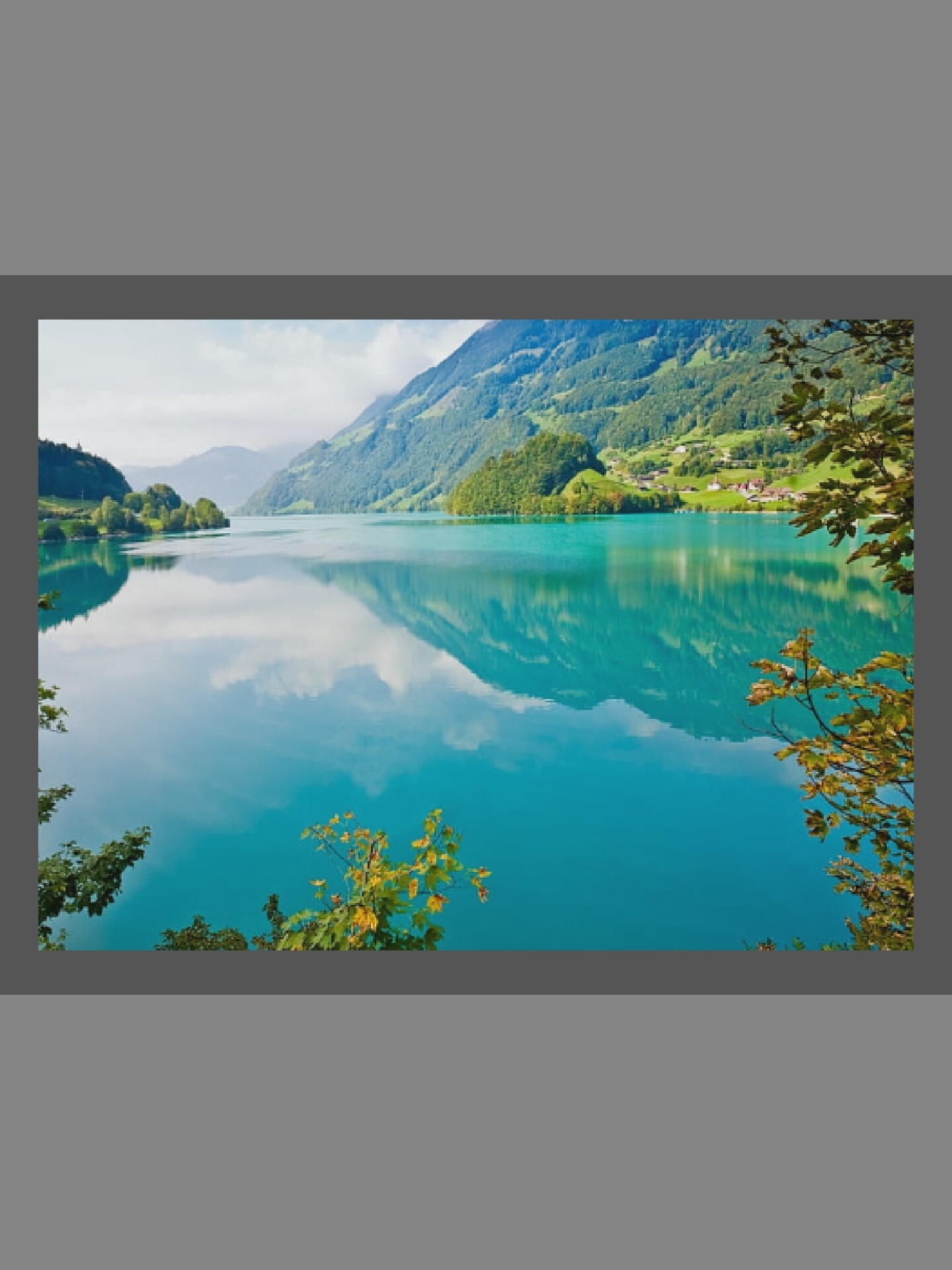 The image displays a serene and majestic landscape featuring a large, vibrantly turquoise lake nestled among lush green mountains. The water perfectly reflects the sky, clouds, and the surrounding forested slopes, creating a striking mirror effect. On the right side of the lake, a small cluster of houses or a village is visible on the lower mountain slopes, integrated into the verdant scenery.
The mountains are steep and densely covered with trees, rising dramatically from the water. Some trees and foliage, with hints of yellow leaves suggesting late summer or early autumn, frame the scene in the foreground, particularly on the right and bottom left. The sky above appears partly cloudy or hazy, contributing to the soft, diffused lighting across the landscape.
The scene conveys a sense of tranquility and natural grandeur. It is daytime, characterized by bright, even illumination. No people or specific activities are depicted, emphasizing the untouched beauty of the environment. The distinct blue-green color of the lake is a prominent feature, often associated with glacial or mineral-rich waters in mountainous regions. No visible text is present. The mountainous terrain and type of lake pictured do not align with the geographical characteristics of Ottawa, Canada.