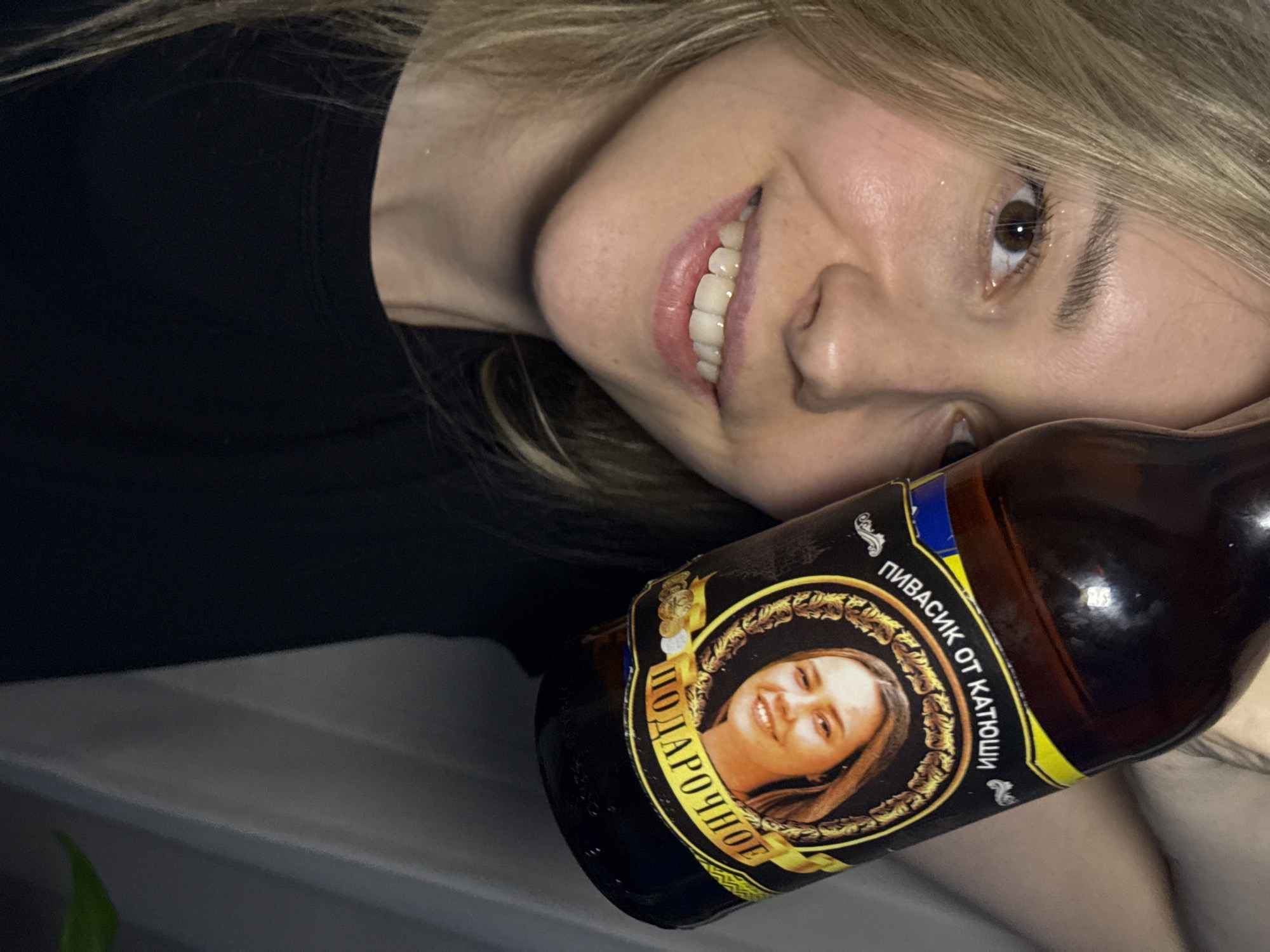 A fair-skinned woman with long blonde hair, smiling, poses closely with a bottle of dark brown liquid, likely beer, displaying the label. The bottle's label includes the words "ПИВАСИК ОТ КАТЮШИ" and "ПОДАРОЧНОЕ," accompanied by a circular ornate frame containing a portrait of a blonde woman. The city of Chaikovsky, Russia, is not discernible from the visual information provided.