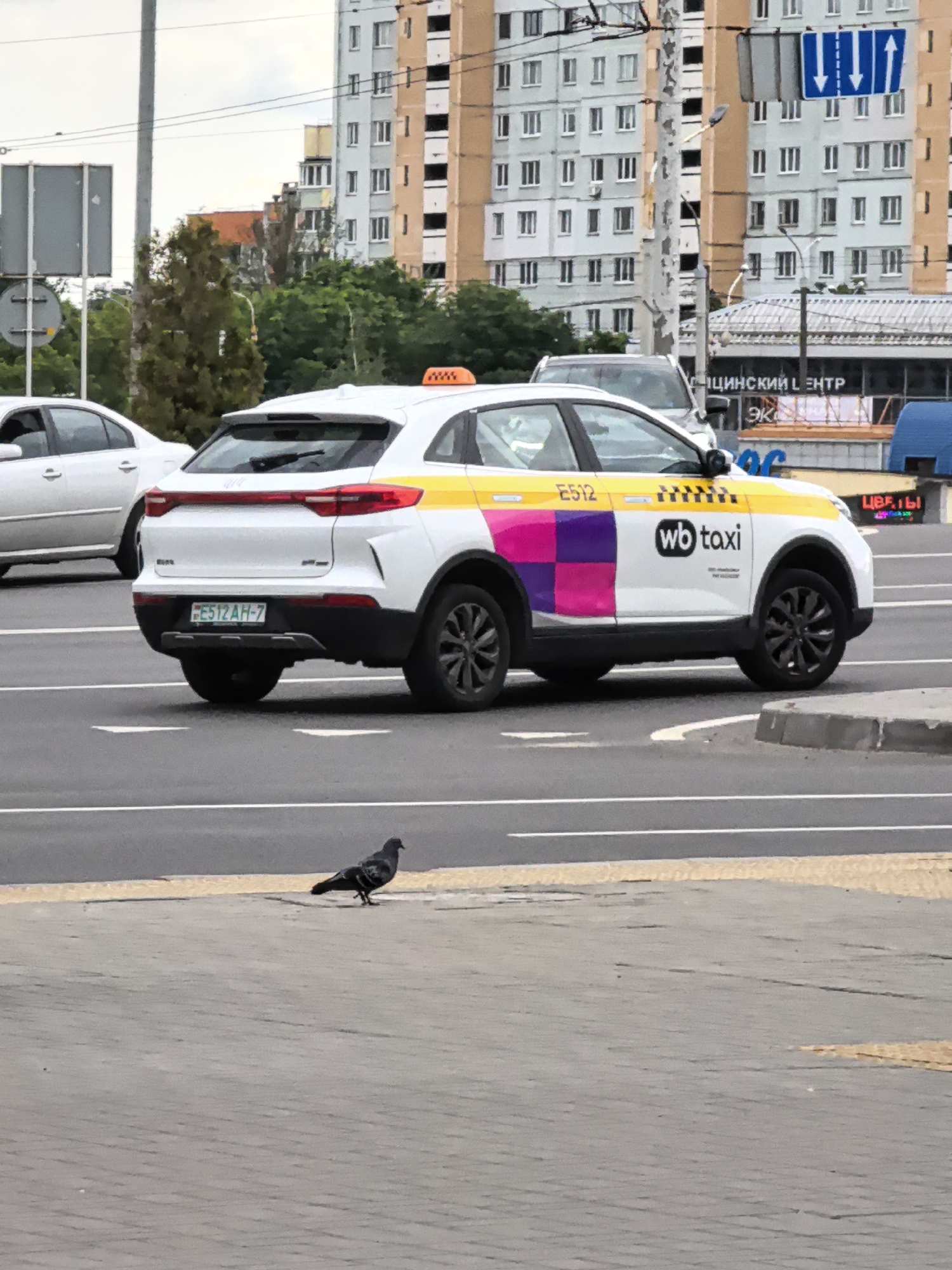 A white taxi with yellow and pink accents is driving on a paved road. The taxi has "wb taxi" written on its side in black lettering. There are buildings in the background, and a traffic light is visible. A pigeon is standing on the sidewalk in the foreground. The weather appears to be overcast. The license plate of the taxi is visible and reads "FE512 AH-7". There is also text on the side of a building in the background, which appears to be in Cyrillic.