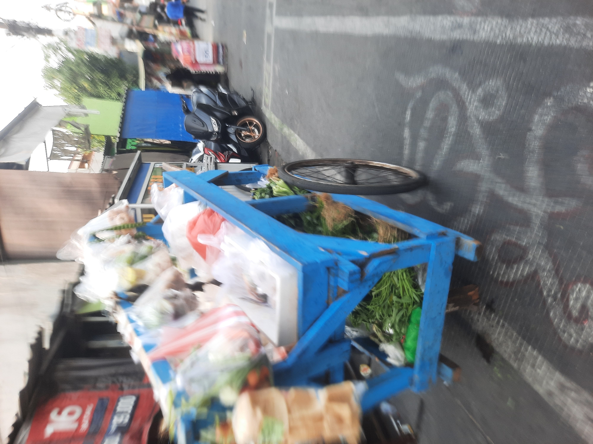 In North Jakarta, Indonesia, an outdoor scene is depicted, likely a street market. A blue wooden cart, filled with produce such as leafy green vegetables and items in plastic bags, is the primary subject. The cart has a wheel on the right side, visible in the foreground. In the background, a black motorcycle is parked, and several people are blurred, moving in the distance. Buildings line the street, one with a red sign displaying the number "16". The overall image is slightly blurred, suggesting movement or low light conditions.