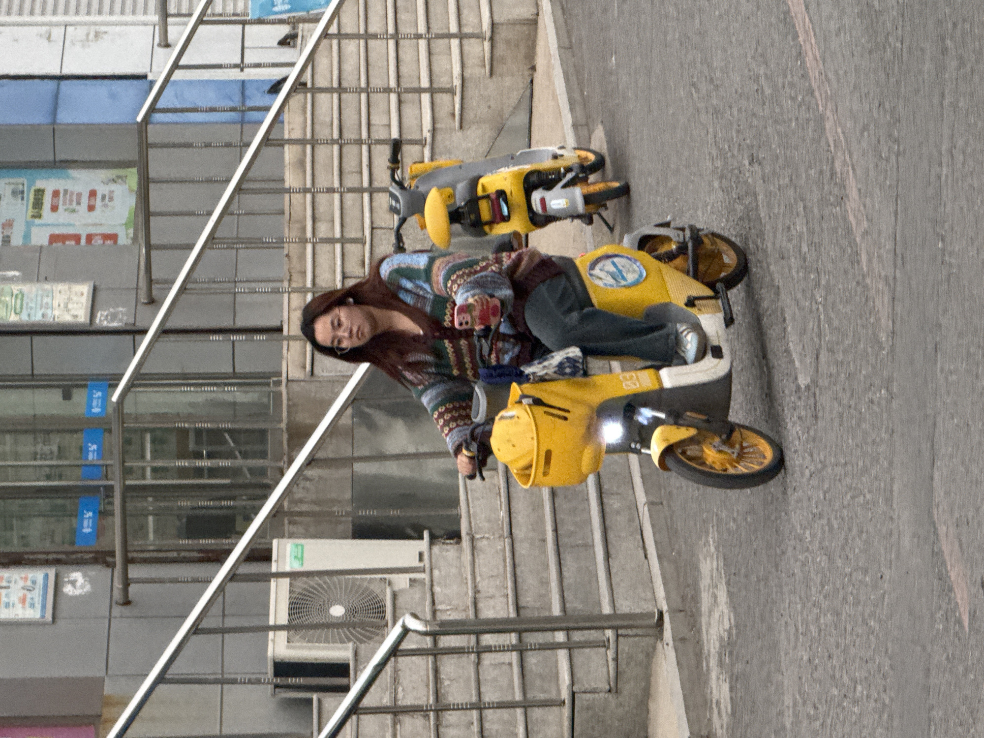 This image, rotated 90 degrees counter-clockwise from its natural orientation, depicts an outdoor urban scene. In the foreground, a young woman with long, dark hair and glasses is seated on a bright yellow electric scooter. She wears a colorful, patterned long-sleeved sweater, dark pants, and white-soled sneakers. Her right hand holds a pink-cased smartphone, which she appears to be looking at or operating.
Another similar yellow and grey electric scooter is parked directly behind the one the woman is on. This second scooter has the number "83" prominently displayed on its front panel. The scooter the woman is using features a blue and white circular logo on its seat, resembling a stylized "N".
The setting includes a paved grey ground or road in the lower right portion of the image. To the left, a set of concrete stairs with silver metal railings ascends upwards. In the background, behind the stairs, is a building facade with glass windows and various signs or posters, some displaying blurry Chinese characters. Blue vertical banners with unreadable text are visible on the building's windows, and a white air conditioning unit is mounted on the wall near the stairs. The lighting suggests it is daytime, possibly overcast, given the lack of strong shadows. The woman's expression is neutral and focused on her phone.