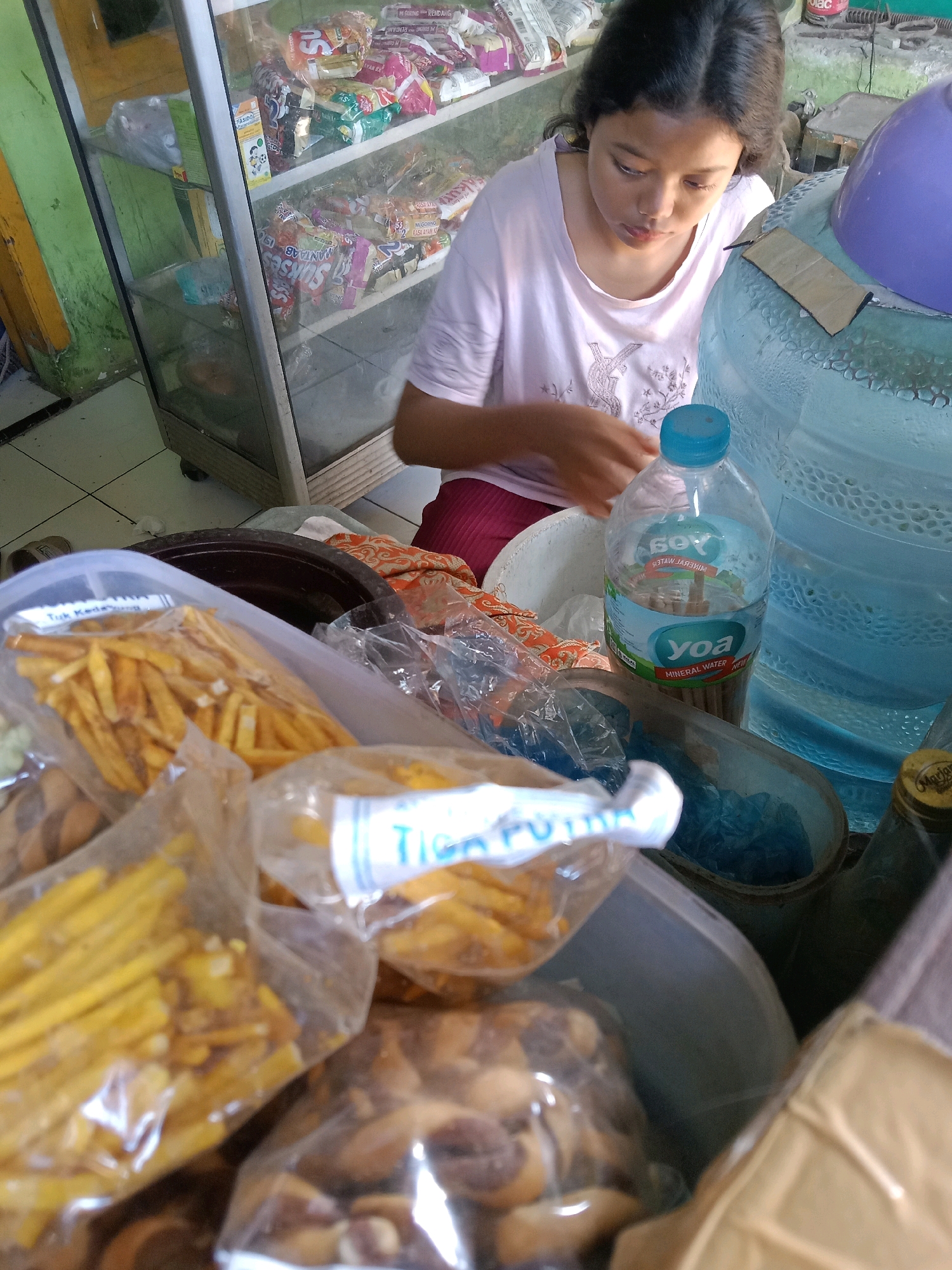 The image captures a young girl in what appears to be a small convenience store or warung in Cirebon, Indonesia. She is wearing a light pink t-shirt and is focused on something in front of her, possibly food items displayed in plastic packaging. She is positioned near a large, clear plastic water dispenser, a common sight in such establishments.
In the background, shelves within a glass display cabinet are filled with various packaged snacks and food items. The environment seems informal and densely stocked, typical of a local shop. Several plastic bags containing different types of snacks, including what look like french fries and other crispy snacks, are prominent in the foreground. A bottle of "Yoa Mineral Water" is also visible, along with other unidentified bottles and containers. The overall lighting suggests an indoor setting with artificial illumination. The image's composition and the presence of food items suggest a scene related to commerce or perhaps preparation of food for sale.