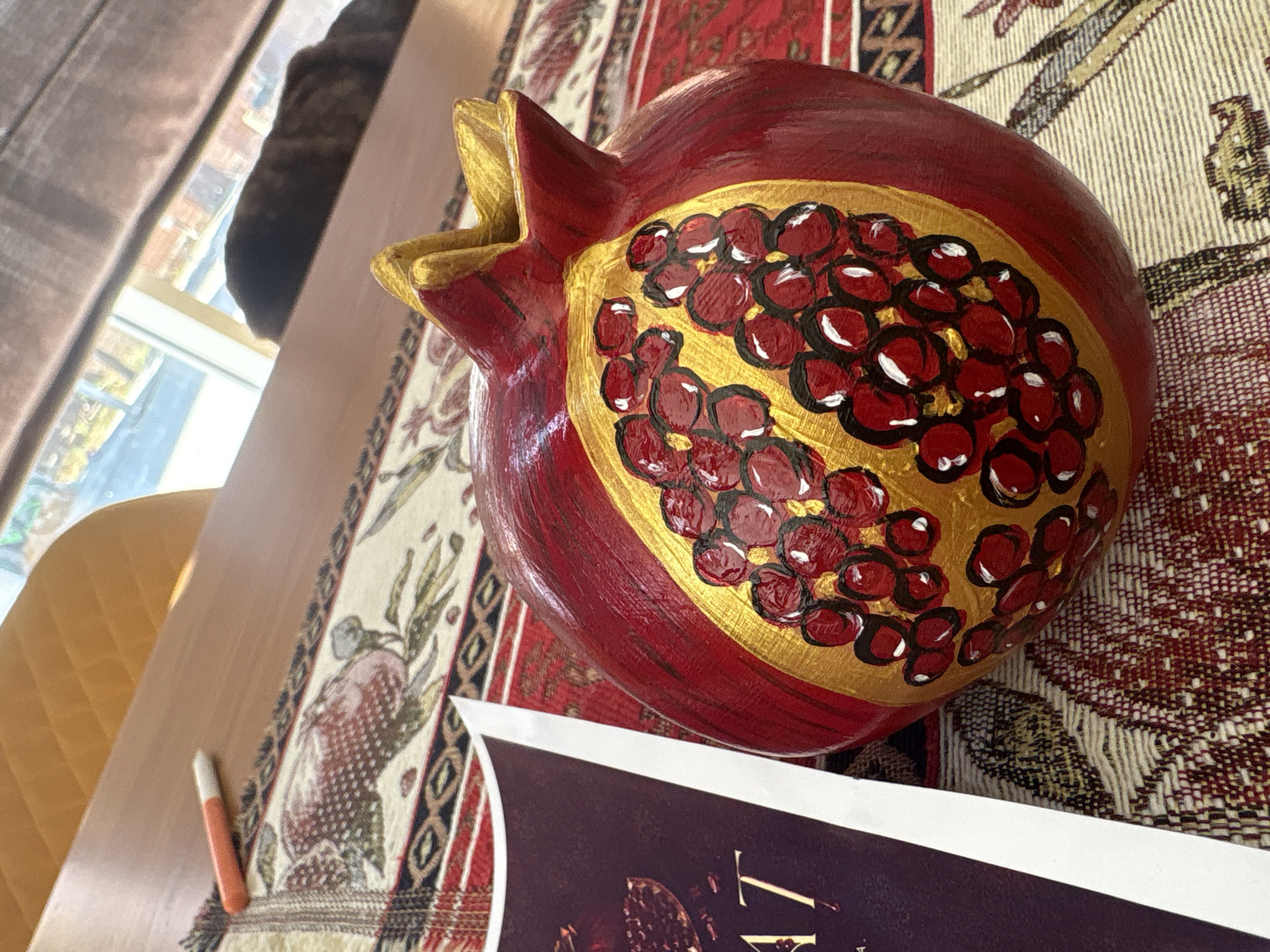 A close-up, eye-level shot showcases a hand-painted decorative pomegranate, predominantly red with gold accents depicting arils, resting on a patterned tablecloth. To the left of the pomegranate, a portion of a booklet with the text "AT" visible is partially covering the tablecloth. The tablecloth itself displays a red and beige abstract floral design. In the background, a wooden table is visible, with an orange-capped marker lying on its surface. Further back, through a window, a dark cat is lying down, its fur blending with the shadows. The environment appears to be indoors, possibly a home setting. The image is captured in Georgiyevsk, Russia.