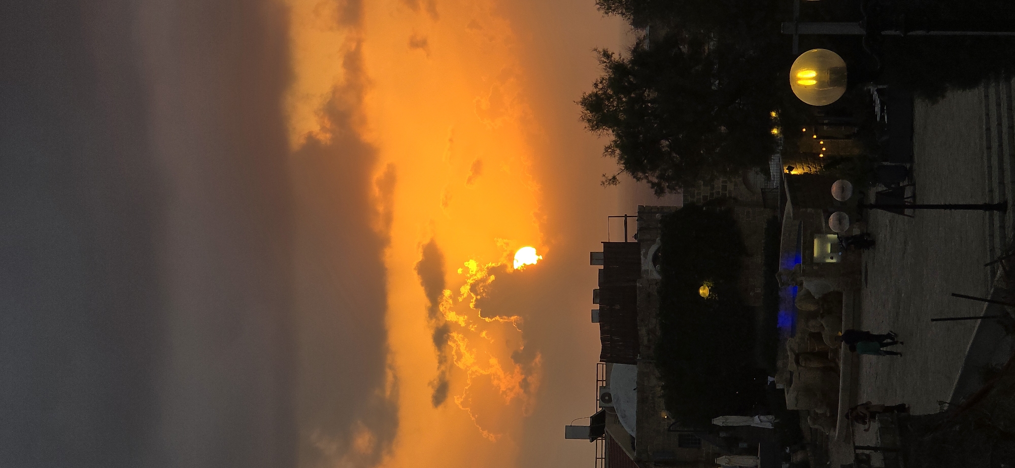 The image captures a sunset over the old city of Jaffa, located south of Tel Aviv, Israel. The sky is dominated by a vibrant orange and yellow hue as the sun, partially obscured by dramatic, dark clouds, dips towards the horizon. In the foreground, traditional stone buildings with flat roofs and architectural elements like arches and battlements are visible. Trees with dark foliage are interspersed among the buildings, casting shadows. A plaza area is visible below, with a few people walking. Streetlights, some with round globes, are illuminated, adding a warm glow to the lower part of the scene, contrasting with the fading daylight. The overall atmosphere is one of a serene evening in a historic coastal city.