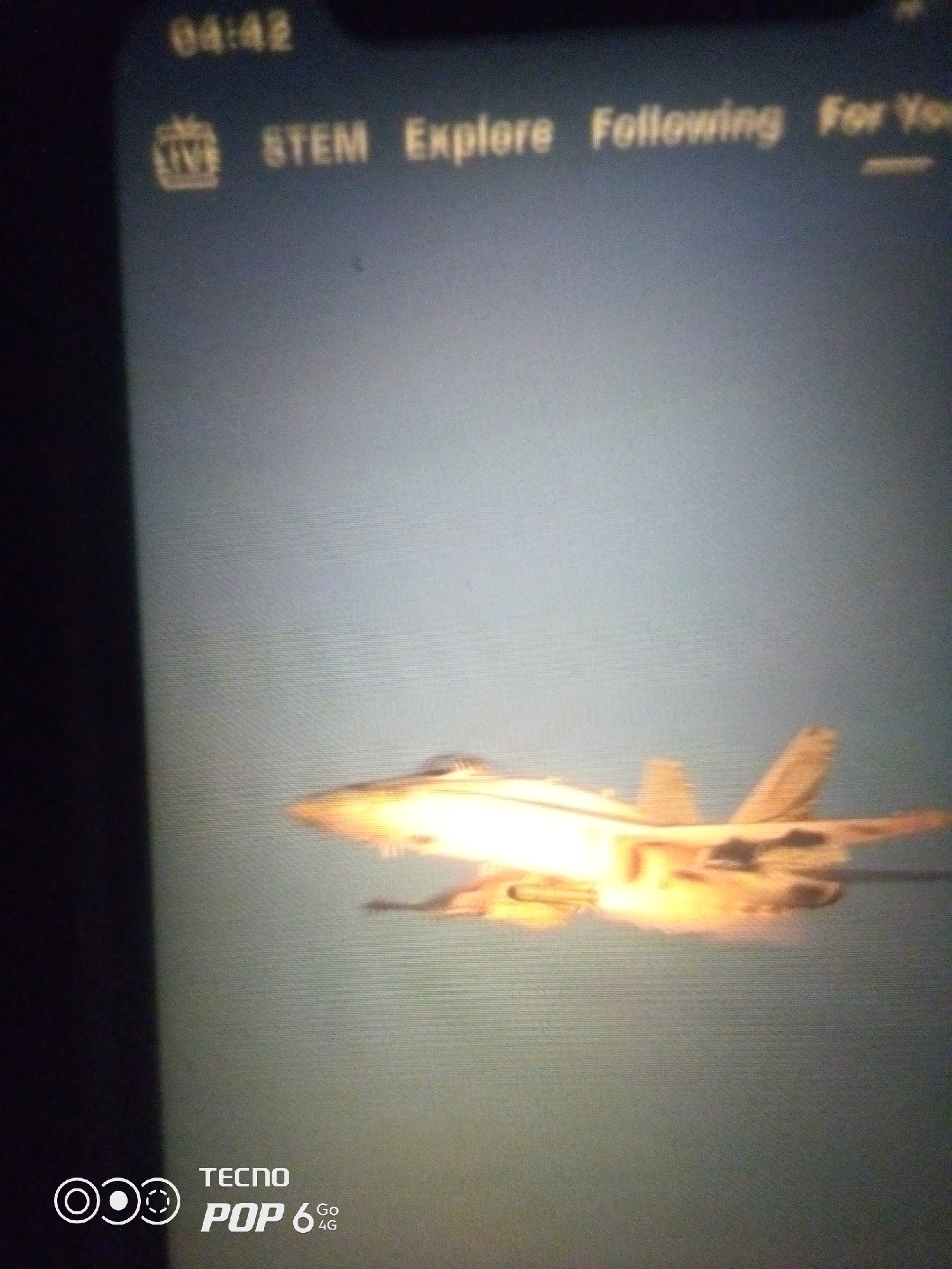 The image displays a phone screen showing a live stream of a fighter jet in flight, with a bright exhaust plume. The top of the screen shows a timestamp of "04:42" and menu options including "LIVE", "STEM", "Explore", "Following", and "For You". In the bottom left corner, the "TECNO POP 6 Go 4G" branding is visible. The background is a pale blue, suggesting it is daytime, possibly over water or a clear sky. There is no visible indication of the specific city of Howayi, Nigeria. The image is a photograph of a phone screen, and the resolution is low, making it difficult to discern fine details.
