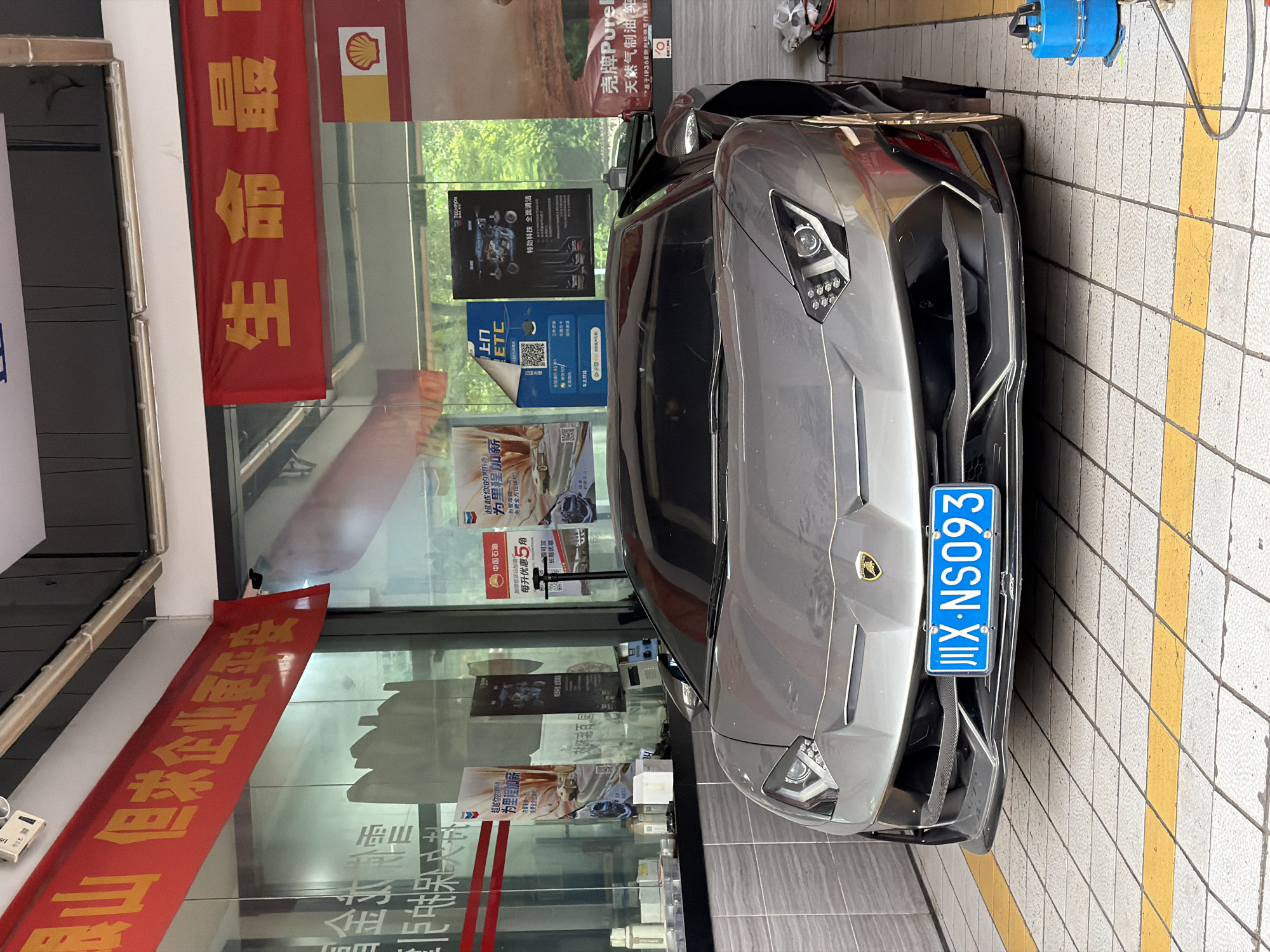 The image captures a grey Lamborghini parked next to a gas station in Chongqing High-tech Zone, China. A Shell sign is prominently displayed on a red banner behind the car. Promotional posters and advertisements are visible on the glass windows of the gas station. The car's license plate reads "川X-NS093".