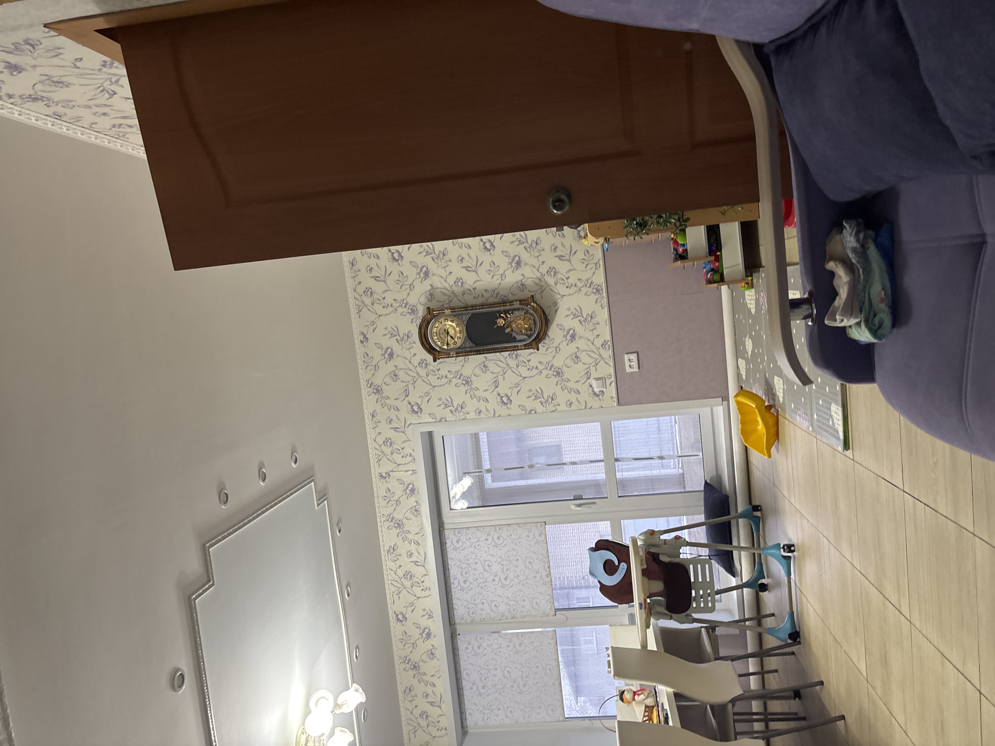 This image depicts the interior of a room in Ферма, Russia. The room's décor includes wallpaper with a floral pattern, a wall-mounted clock, a chandelier, and a mirror with decorative accents. A brown wooden door is prominent in the upper part of the image. A partially visible blue sofa is in the upper right. The room appears to have a window with blinds and features a high chair with a brown seat, and what seems to be a dining area with light-colored chairs. A yellow plastic container is on the floor. There is a baby swing with clothing hanging on it. The walls are light gray.
