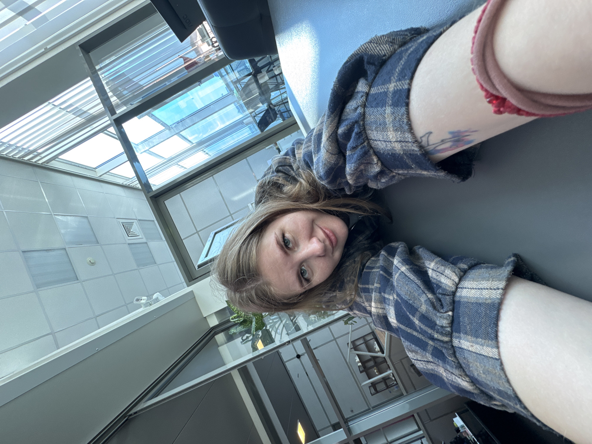 The media file is a diagonally oriented selfie taken by a young woman in a brightly lit, modern indoor environment. The woman, with light brown hair and blue eyes, is looking directly at the camera with a relaxed, soft smile. She is wearing a blue and beige plaid flannel shirt. On her visible right arm, she has a light-colored wristband or scrunchie and a small, colorful tattoo, appearing to be a floral or abstract design in pink and blue hues.
The background reveals a contemporary architectural space, characterized by an expansive skylight system made of glass panels and metal frames. This structure allows significant natural light to illuminate the interior, suggesting it is daytime with a clear sky visible through the glass. The setting appears to be a public or commercial building, such as an airport terminal, a modern shopping center, or a large corporate lobby.
Further details in the background include white ceiling tiles with visible ventilation grilles and a fire alarm/smoke detector. A white, dual-lens security camera is mounted on a ledge, reinforcing the public nature of the location. Reflections on the glass surfaces show distorted views of the interior, including some green foliage and a dark object bearing the text "AOC," likely a brand name for a monitor or display. The overall impression is one of a clean, spacious, and architecturally designed interior, consistent with modern facilities found in cities like Perm, Russia.