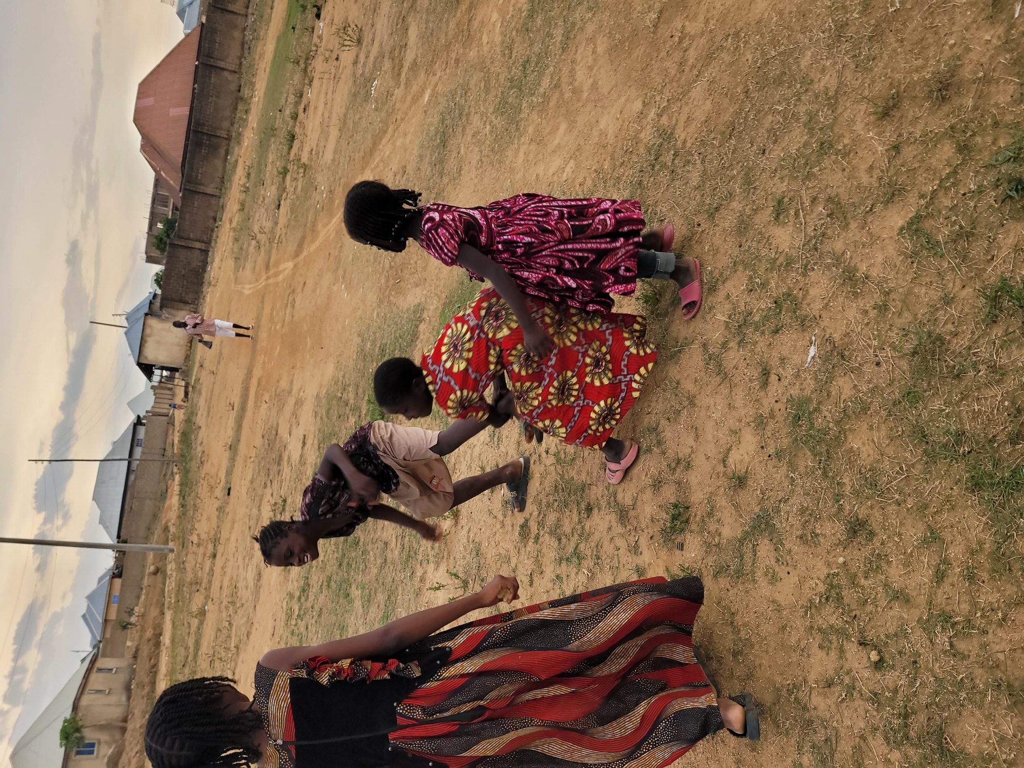 The photograph shows a group of four young girls playing in a dirt field in what appears to be Barakin Akawo, Nigeria. Three of the girls are interacting closely, in what appears to be a game of some kind. One girl wears a red and white patterned dress, another a red floral pattern dress, and the third a tan dress. The fourth girl is standing, wearing a red and black dress. In the background are structures indicative of a residential area, including houses with corrugated metal roofs and a high wall. The sky is overcast.