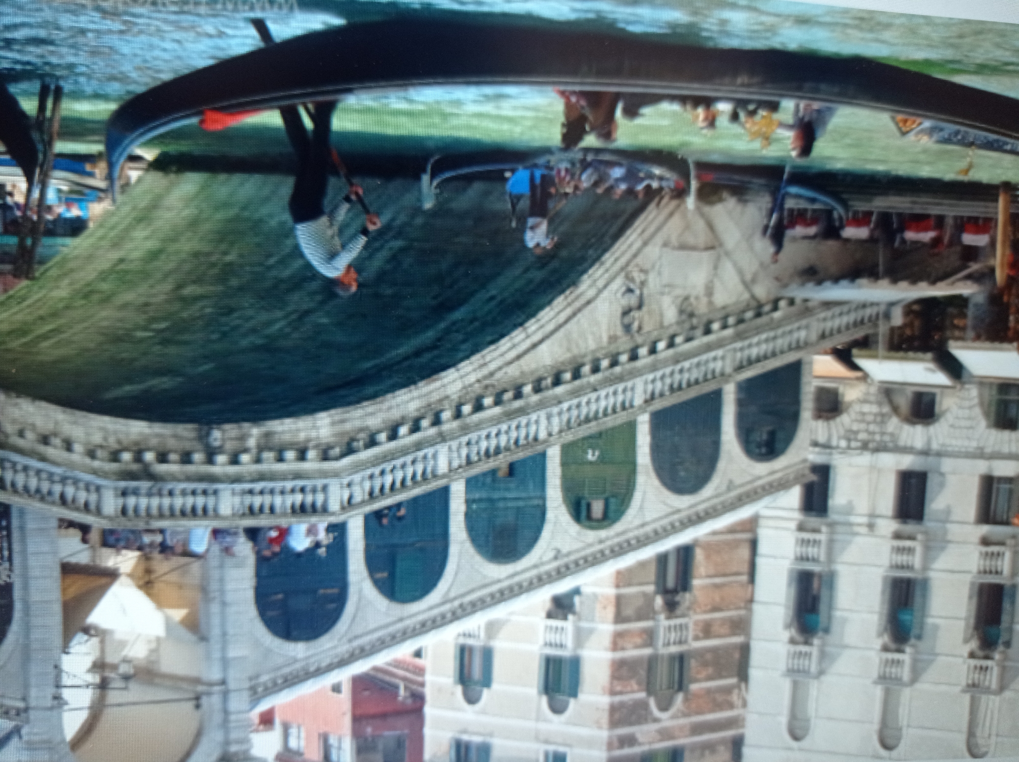 This is a photograph of the Rialto Bridge in Venice, Italy, not Mukachevo, Ukraine. The image captures the iconic bridge spanning a canal, with gondolas navigating the water below.
On the bridge, people are visible walking and standing along the railings, with some appearing to be tourists. Shops or restaurants with red-and-white striped awnings are situated on the ground level of the bridge's structure.
In the foreground, several gondolas are present. One gondola in the immediate foreground is occupied by a person in the front, likely a gondolier, steering with a single oar. In another gondola further back, multiple passengers are seated. A gondolier in a striped shirt and dark pants is actively rowing a gondola towards the right side of the frame. The water is a murky green, reflecting the surrounding architecture and sky. The buildings on either side of the canal are typical Venetian architecture, with ornate facades and arched windows. The overall scene depicts a bustling and picturesque day in Venice.