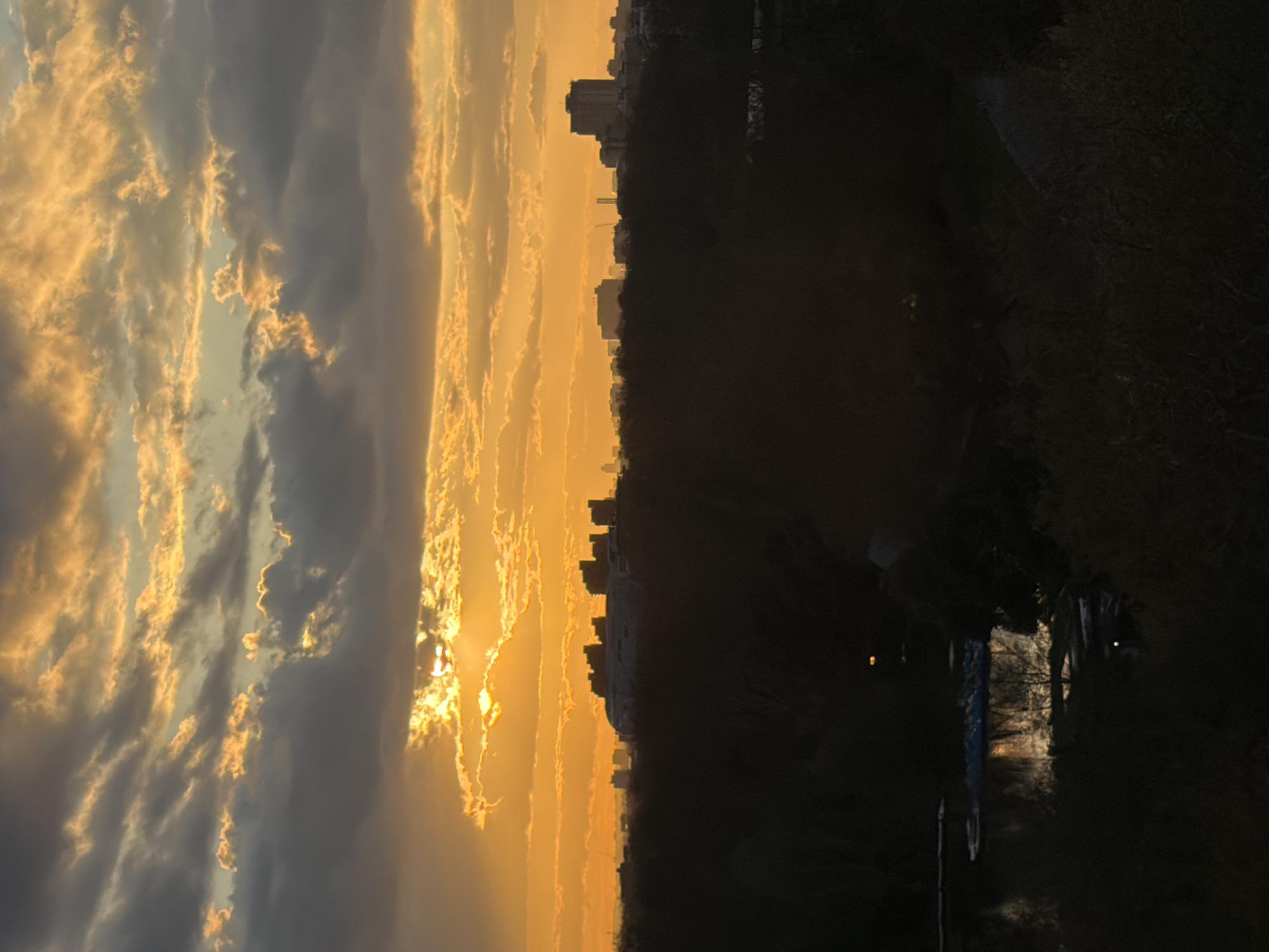 The image shows a cityscape at sunset. In the foreground, a dark, shadowed area with trees and possibly a body of water is visible. The middle ground features a city skyline silhouetted against the setting sun. The sky is filled with golden and grey clouds, with the sun's rays breaking through in some areas. The image is taken at eye level. The photograph is taken from a location along the edge of Regent's Park. The skyline shows buildings along the north west portion of London.