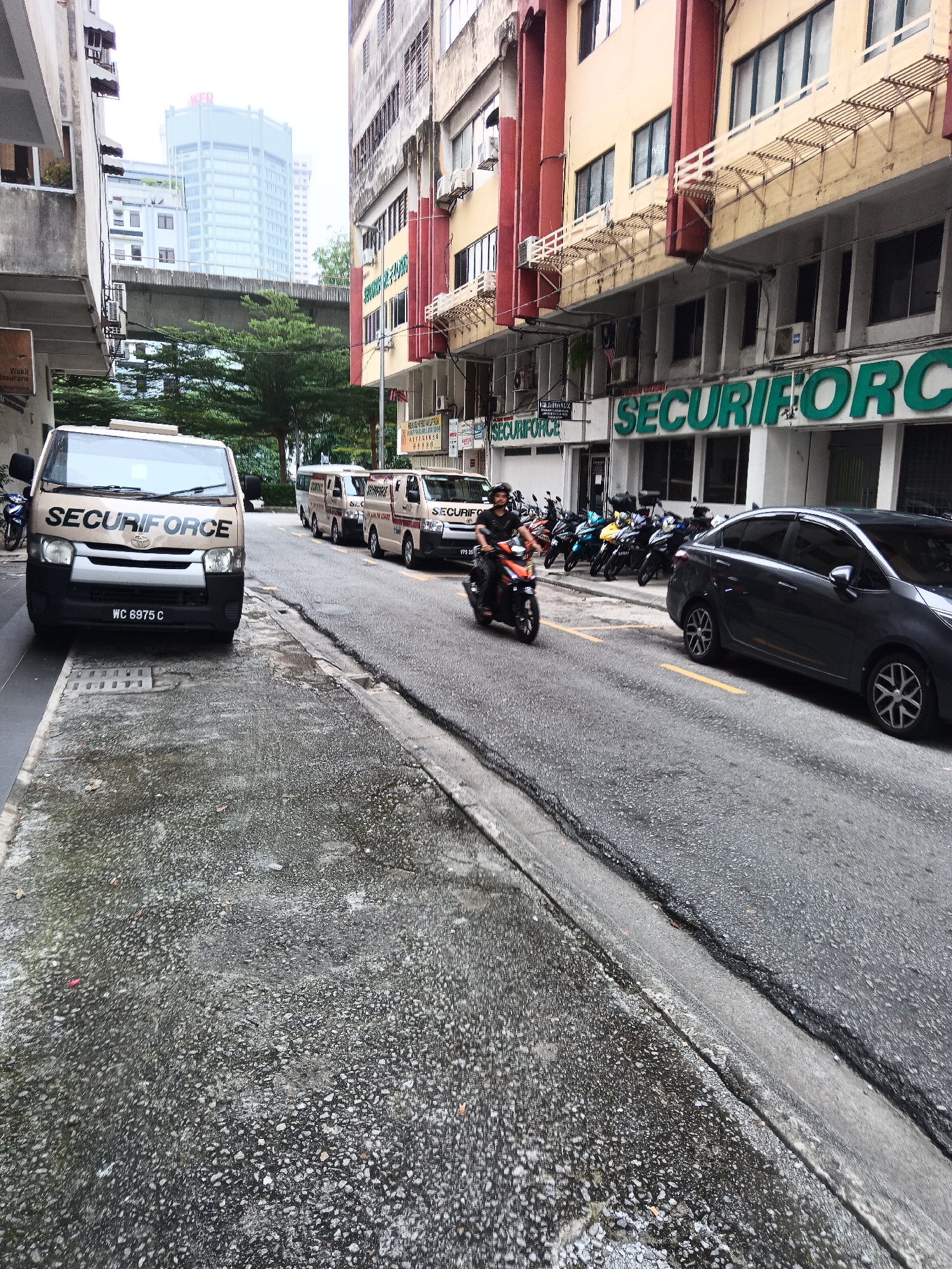 In Kuala Lumpur, Malaysia, an urban street scene unfolds, featuring a line of securiforce armored vans and motorcycles parked along the roadside. The buildings are multi-story, with visible air conditioning units and balconies. The street itself is paved, with a curb separating it from the sidewalk. A dark sedan is parked on the street, and a man is riding a motorcycle. The lighting suggests an overcast day.