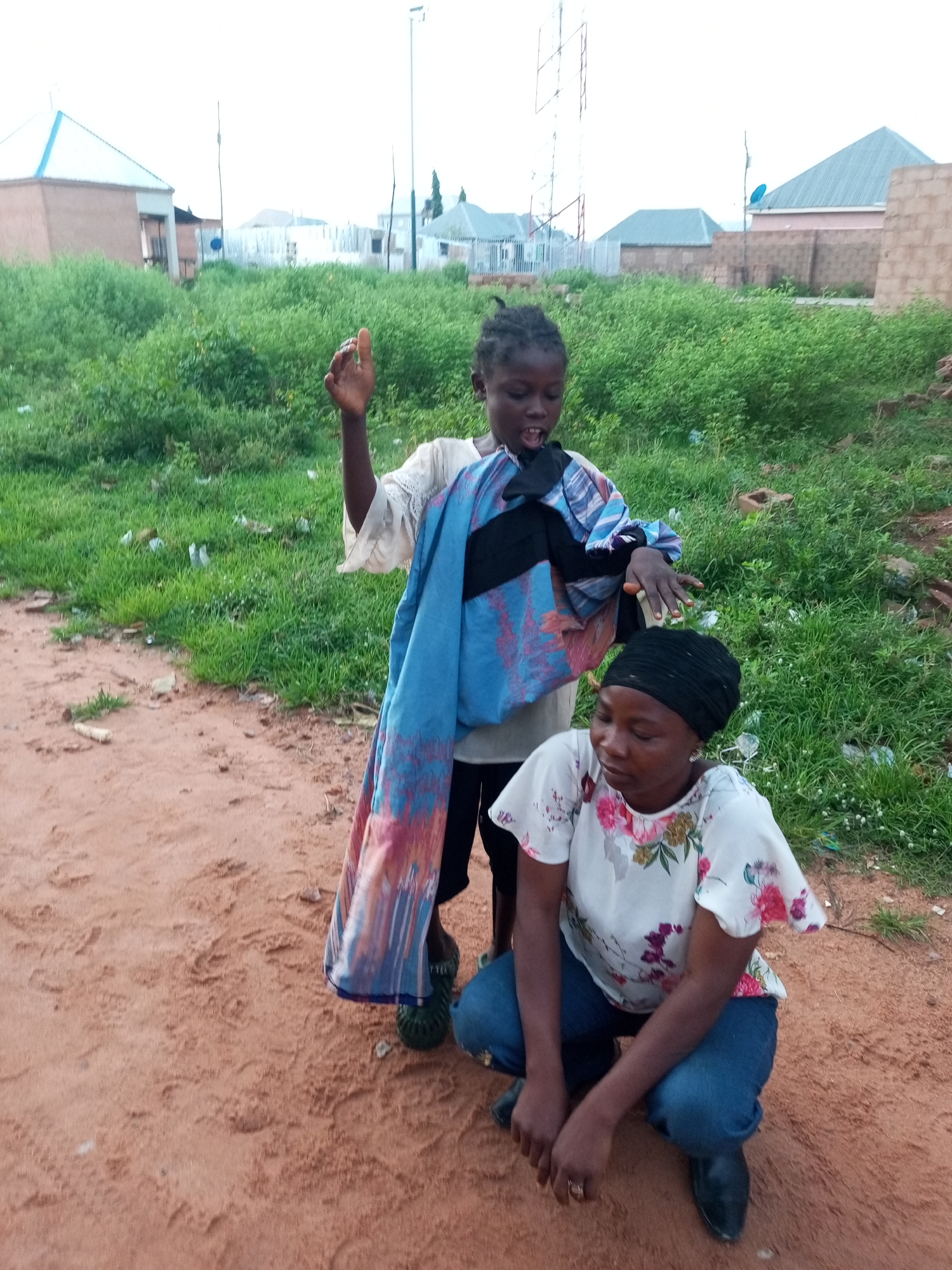 The image depicts two individuals in an outdoor urban environment, likely in Tempure, Nigeria. A young girl, standing, holds a bundle of clothing, primarily blue and pink patterned fabric, draped over her arm and shoulder. She wears a light-colored, possibly cream or beige, embroidered top and dark trousers. Her mouth is open as if speaking or singing, and her right hand is raised in a gesture. Beside her, a woman is crouched down. She wears a black headscarf and a white short-sleeved shirt with a floral print of pink and green flowers. Her lower body is clad in blue jeans. Both individuals are positioned on a reddish-brown dirt path. The background consists of overgrown green grass, some scattered debris, and in the distance, various buildings with grey and light blue roofs, along with a telecommunications tower. The sky is overcast.
