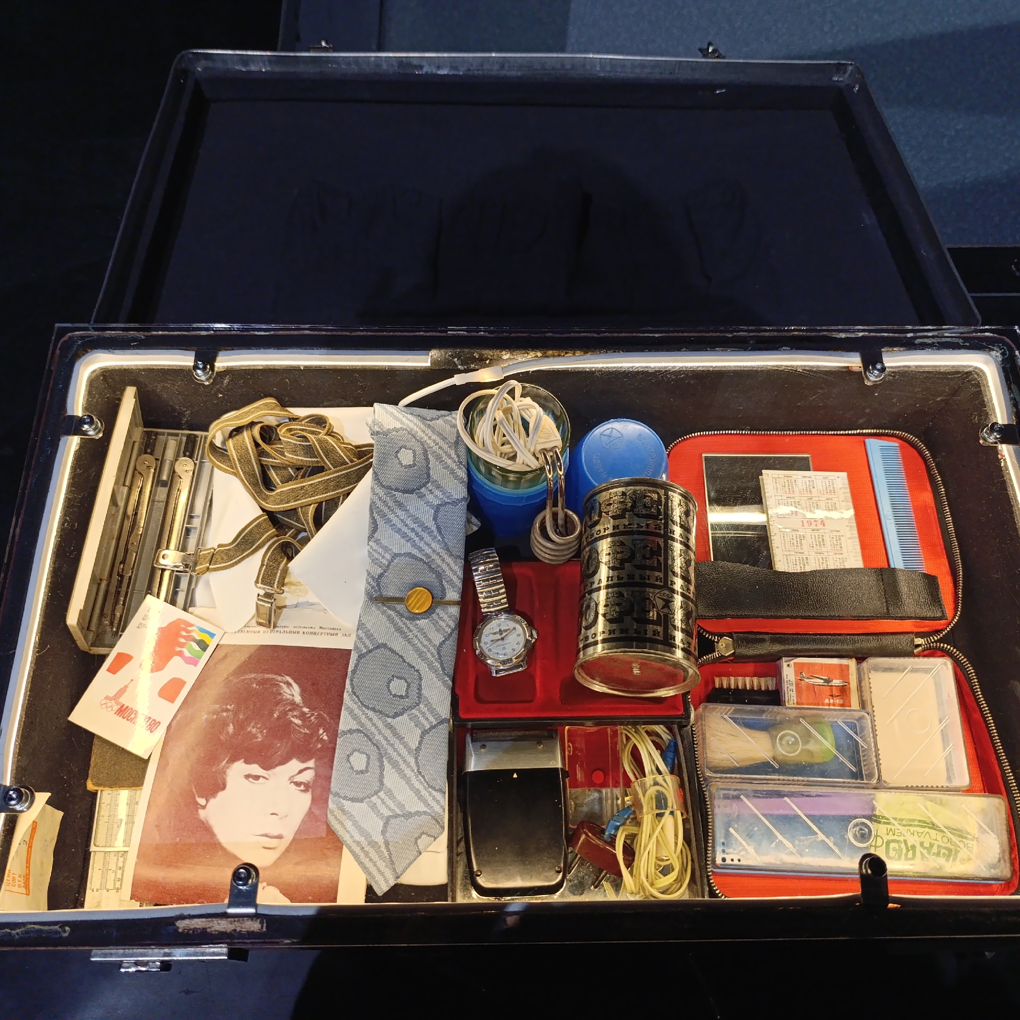 This image captures an open black briefcase or trunk densely packed with a variety of personal items, likely dating from the Soviet era. The dark interior of the case and its lid serve as the background for the collection.
The contents of the case include items for personal care and travel, arranged somewhat haphazardly yet fittingly. On the left, a pair of brown and white suspenders are coiled next to various papers. These papers include a black and white photograph of a woman with short, dark hair, a flyer related to the "Moscow 80" Olympics, and a program or document from the "Московский Центральный Концертный Зал" (Moscow Central Concert Hall).
Towards the center, a light blue patterned tie is folded, alongside a silver-toned wristwatch. Nearby are a set of blue and green plastic stacking cups, with a coiled immersion heater and its electrical cord suggesting preparation for making hot beverages while traveling. A tall, cylindrical metal container, bearing Russian text and "СССР" (USSR), stands prominently.
On the right side of the case is a red zippered travel bag containing a mirror, a light blue comb, and smaller transparent plastic containers. One of these containers holds a brush, another contains a matchbox with an airplane design, and another has visible text including "BOROTVAKREM," likely a brand name for shaving cream. A small calendar marked "1974" is also visible within this section.
The ensemble of objects, particularly the 1974 calendar, "Moscow 80" reference, and Soviet markings, strongly indicates a time frame between the 1970s and early 1980s. The assortment suggests a personal travel kit or a collection of everyday items from that period, possibly displayed as part of a historical exhibit in Verkhnyaya Pyshma, Russia. No direct activity is depicted; it is a still life showcasing a moment in time through personal belongings.