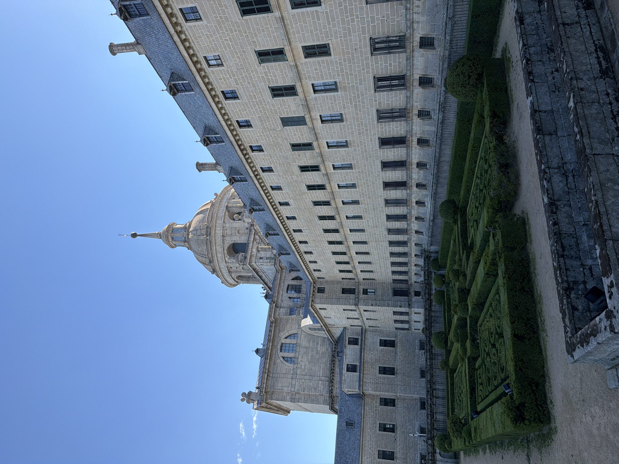The image shows a view of the Royal Site of San Lorenzo de El Escorial in San Lorenzo de El Escorial, Spain. The photograph captures a portion of the massive complex, highlighting its architectural grandeur. In the foreground, a meticulously manicured garden with intricate hedge mazes and rounded topiary bushes is visible. Beyond the garden, the imposing stone facade of the monastery and palace stretches across the frame, characterized by rows of regularly spaced windows with dark shutters and small dormer windows on the steeply pitched slate roof. A prominent dome with a spire crowns a section of the building in the background, silhouetted against a clear blue sky. The stone construction of the building is detailed, with visible mortar lines between the individual blocks. The lighting suggests it is daytime, likely morning or afternoon, with the sun casting soft shadows. There are no people visible in the image. The overall impression is one of historical significance and architectural symmetry.