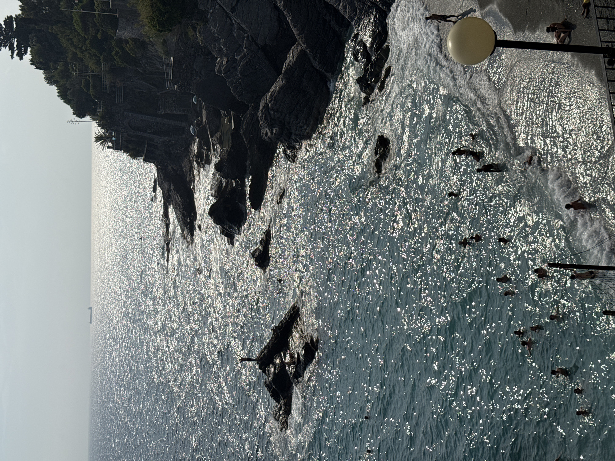 The image captures a sun-drenched view of the Ligurian Sea in Bogliasco, Italy. The sea is a vibrant blue, reflecting the bright sunlight with a shimmering, almost sparkling effect. Along the right side of the frame, a rocky coastline is visible, covered in lush green vegetation. Dark, rugged rocks jut out into the water, creating small coves and inlets.
In the foreground, a sandy beach transitions into the sea, where numerous people are enjoying the water. Some are swimming, while others are wading near the shore. The waves are gentle, creating a foamy white edge as they meet the sand. A single, tall lamppost stands on the beach, its large spherical light fixture glowing softly. Several people are seated or lying on the sand near the lamppost, appearing to relax.
Further out in the sea, a lone figure stands on a dark rock, arms outstretched, silhouetted against the bright expanse of water. In the far distance, a large cargo ship is visible on the horizon, a small dark shape against the bright sky. The overall scene conveys a sense of a peaceful and lively summer day at the seaside.