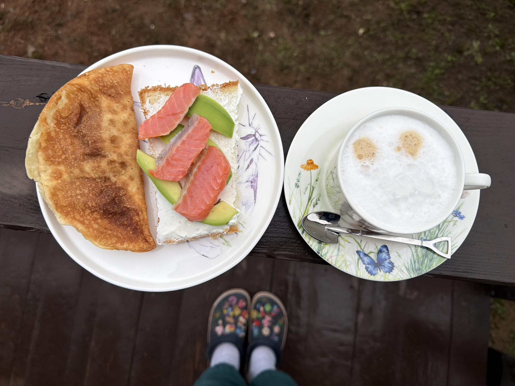 The image presents a top-down view of a meal and beverage arrangement, accompanied by a partial view of a person's feet. The meal consists of a plate with a portion of flatbread and an open-faced sandwich composed of bread, cream cheese, avocado slices, and smoked salmon. Adjacent to the plate is a cup of frothy beverage, likely coffee or tea, sitting on a saucer. Two spoons rest beside the cup.
Below the plates and the beverage is a person's feet wearing dark-colored shoes with multicolored floral decorations. The setting appears to be outdoors, possibly on a wooden deck or porch, with blurred greenery visible in the background.
The exact location within Chaikovsky, Russia, cannot be determined from the visual data alone.