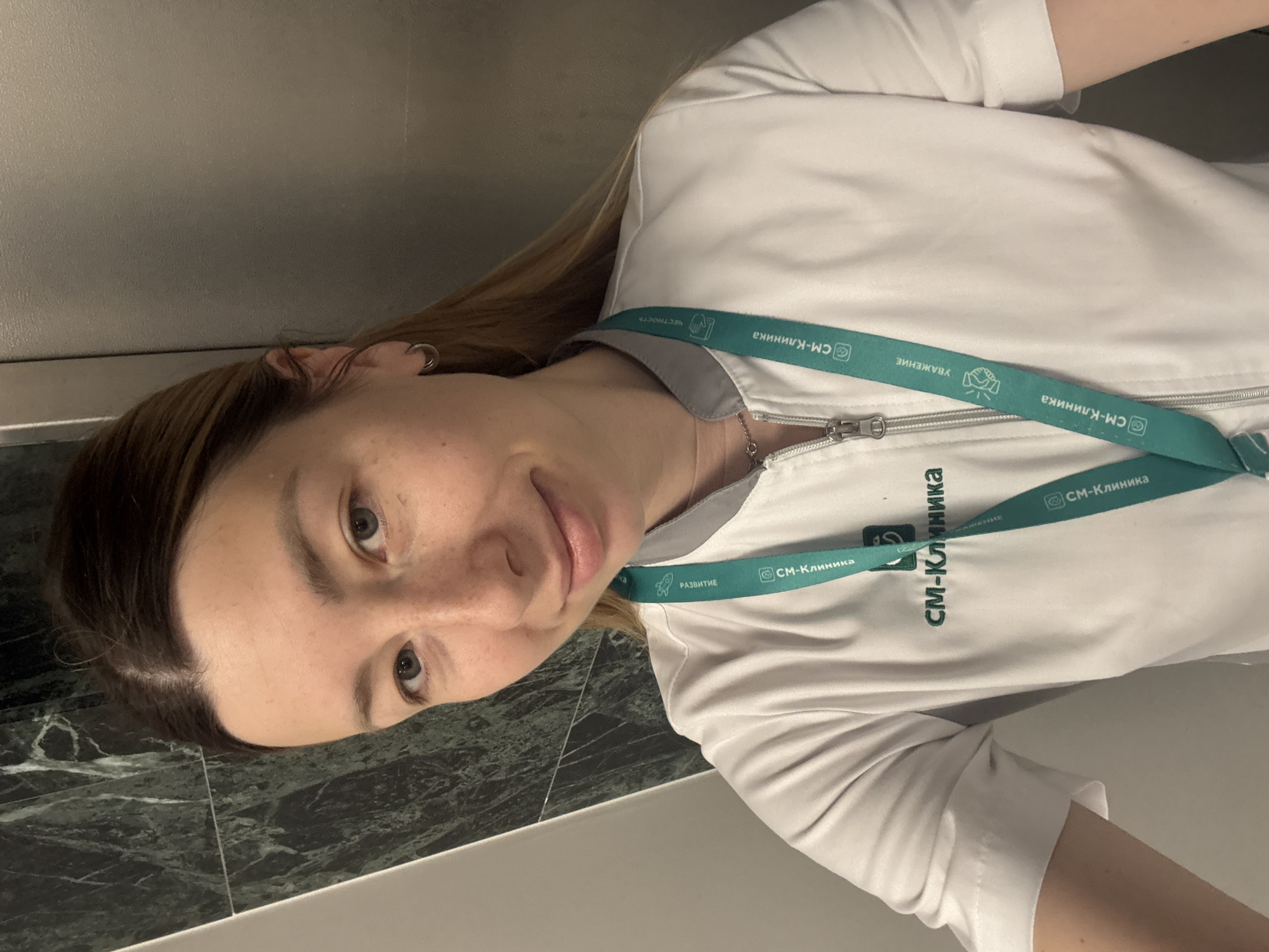A woman in a white medical uniform is in an elevator. She is wearing a lanyard with the "SM-Klinika" logo and several Russian words, including "РАЗВИТИЕ" (Development), "ЧЕСТНОСТЬ" (Honesty), and "УВАЖЕНИЕ" (Respect). She has blonde hair, light eyes, and is smiling. The elevator walls are metallic gray and dark green marble. The location is not specified in the image.