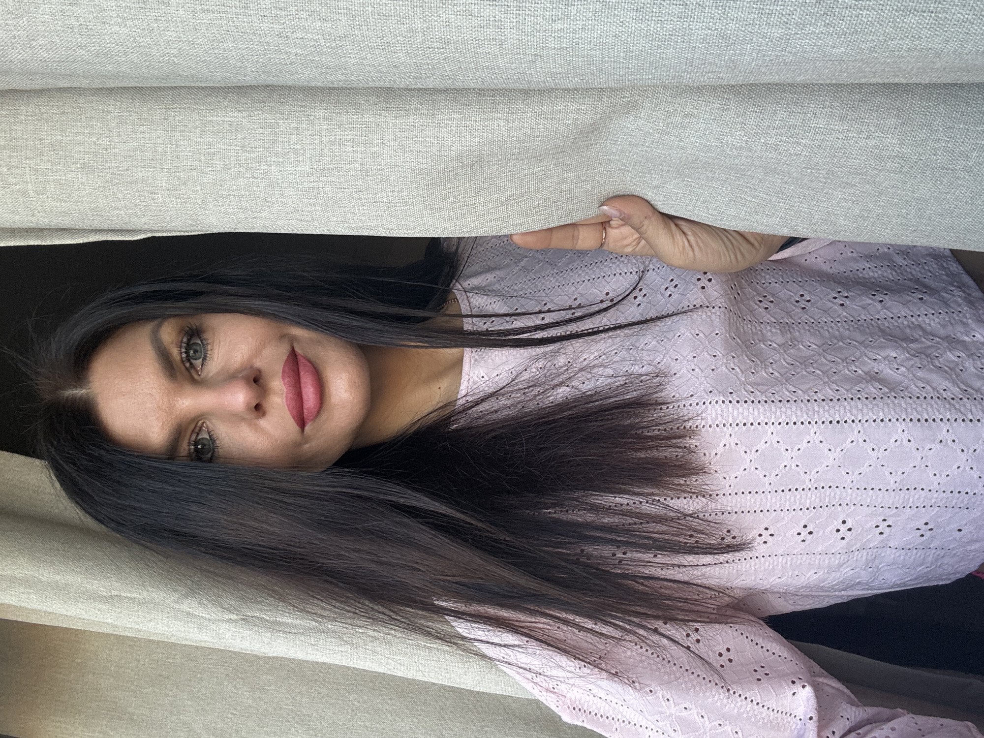 A woman with long, dark hair and striking green eyes peeks playfully from behind two light-colored curtains. She has pink lipstick and offers a soft smile directly at the viewer. Her head is slightly tilted. Her right hand, adorned with a simple ring on her ring finger, is visible as she holds the textured, light beige or gray curtain aside, revealing her. She is wearing a light pink blouse featuring an intricate eyelet or embroidered pattern. The background behind the woman appears dark and indistinct, suggesting an interior space or the shadows beyond a window. The lighting appears natural, indicating it is daytime. No text is visible in the image. The overall impression is one of curiosity or a friendly greeting.