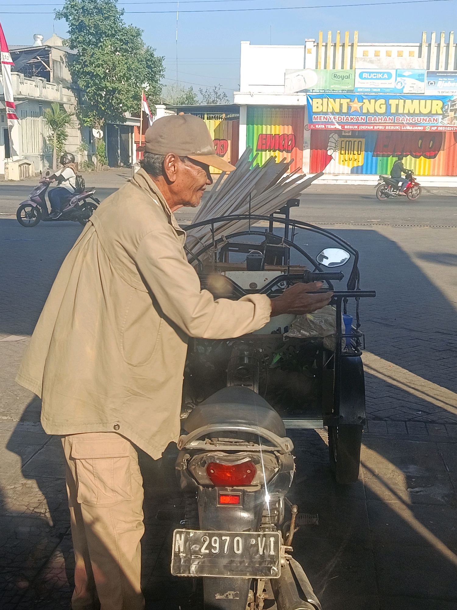 The image depicts a street scene in Tempeh Lor, Indonesia, under a clear sky. In the foreground, an elderly man wearing a light brown cap and a tan jacket and pants stands next to a motorcycle with a trailer. He is holding onto the handlebar of the trailer, which is filled with long, thin wooden planks. The motorcycle's license plate reads "N 2970 VI." To the left of the man, a purple scooter with a rider passes by. In the background, a building with a colorful mural and various signs, including "BINTANG TIMUR," is visible. Further down the street, another person rides a red motorcycle. The ground is paved with cobblestones and asphalt, and shadows from the man, the motorcycle, and other unseen objects are cast on the ground.