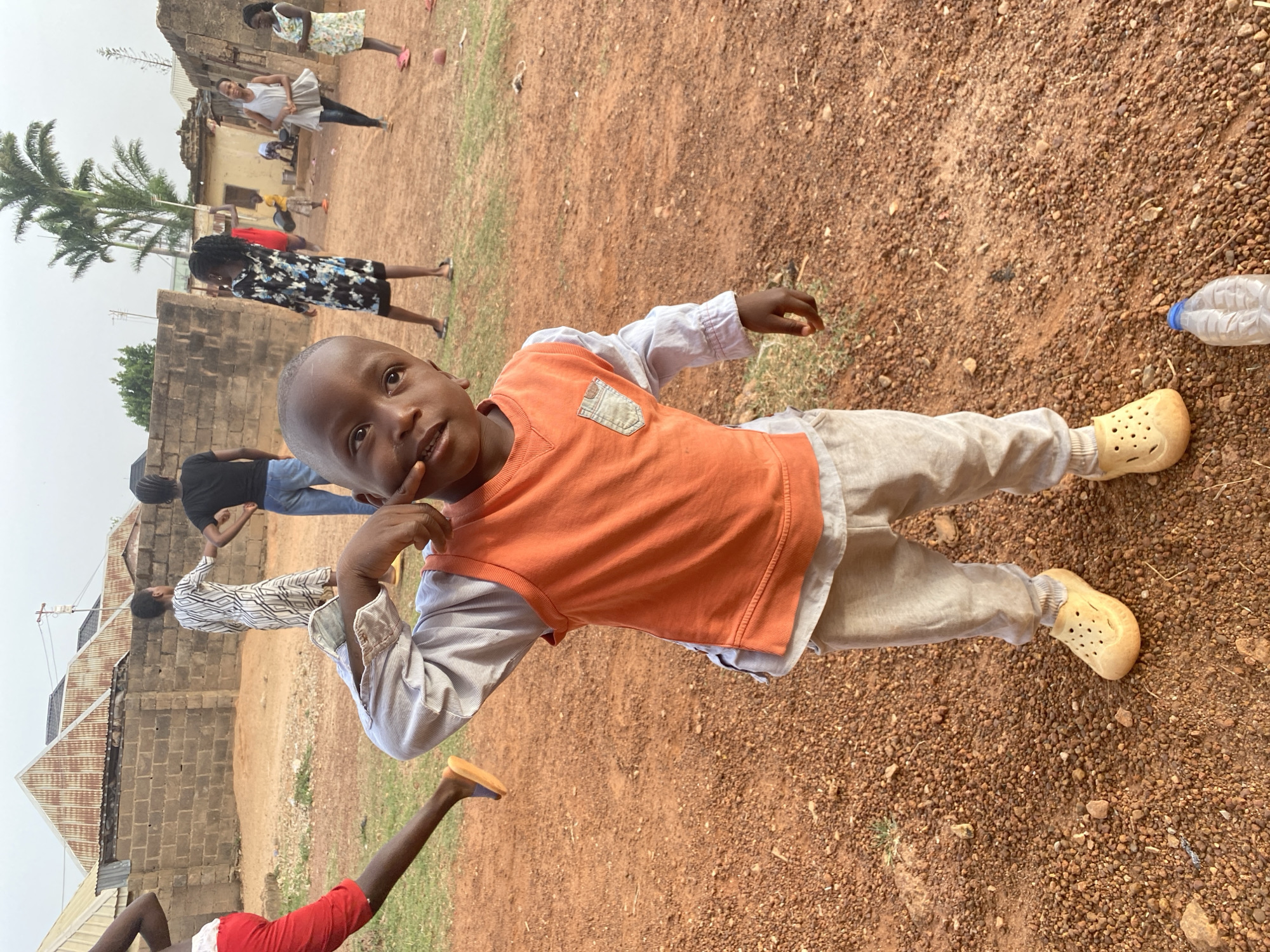 The image captures a full-length shot of a young Black boy in an outdoor setting in Bukuru, Nigeria, looking directly at the camera with a curious or slight smiling expression. He is wearing an orange sleeveless top over a light-colored long-sleeved shirt, light tan trousers, and light yellow/beige clogs. His right index finger is resting near his mouth.
The ground is a reddish-brown, stony earth characteristic of the region. In the mid-ground and background, several other Black individuals are visible. These include a woman in a black and white patterned dress, another woman in a white top and dark bottoms, and other partially obscured figures, some appearing to be children, in casual clothing. Buildings with exposed brick walls and corrugated iron roofs are visible in the background, along with a tall palm tree on the left side of the frame. A clear plastic water bottle lies on the ground to the right of the main subject. The scene is brightly lit, indicating it is daytime, likely midday or early afternoon, under sunny or lightly overcast conditions. The overall atmosphere appears casual and relaxed, depicting an everyday outdoor scene. No visible text is present in the image.