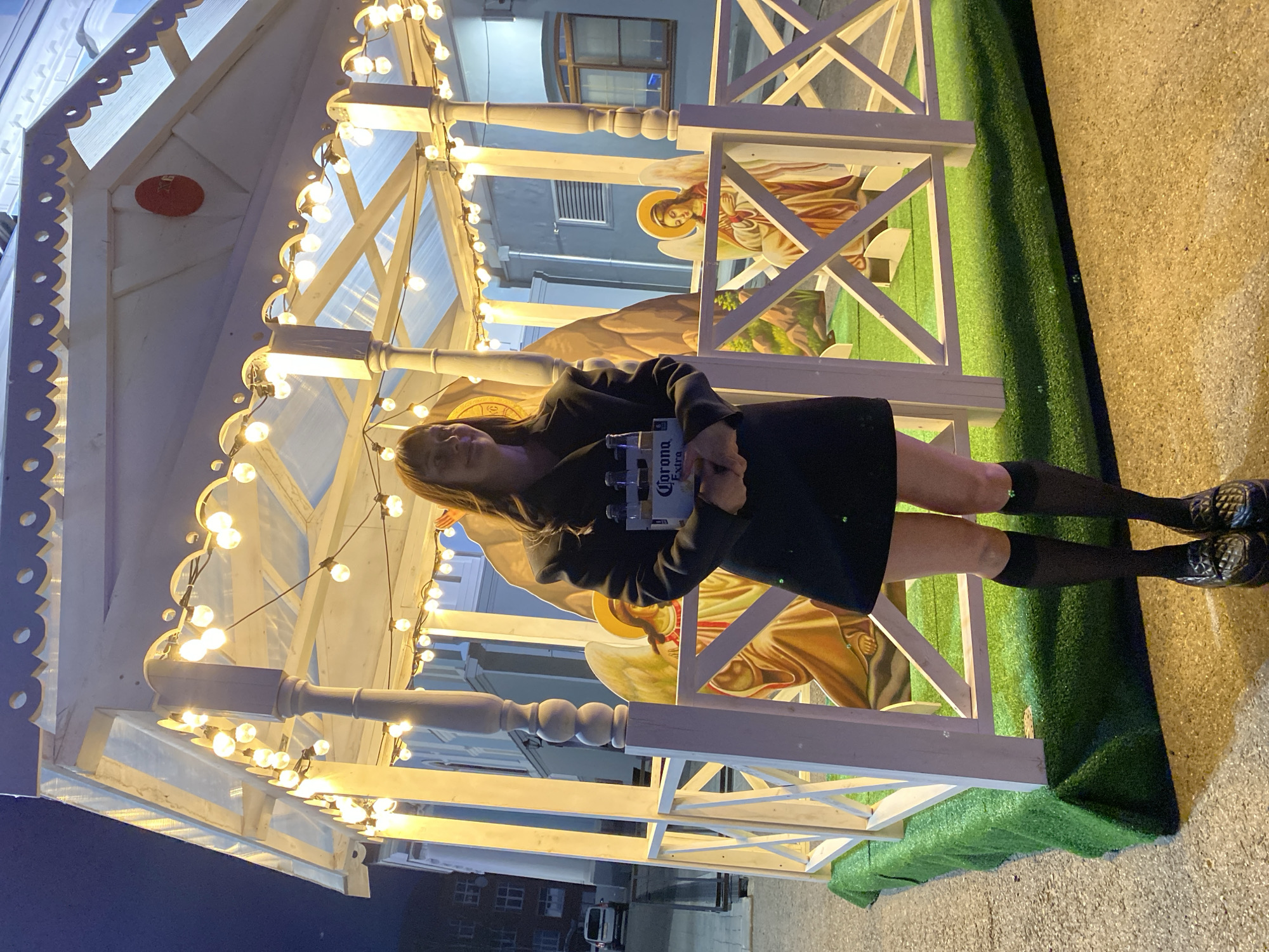 A woman holds a six-pack of Corona Extra beer in front of a Christmas nativity scene replica in Votkinsk, Russia. The scene, under a wooden A-frame structure adorned with strings of lights, features figures of angels. The woman, wearing a black dress and knee-high socks, faces the camera. The exterior lighting is low. The scene is set on a concrete surface with a patch of artificial grass. Nearby, streetlights illuminate surrounding buildings.