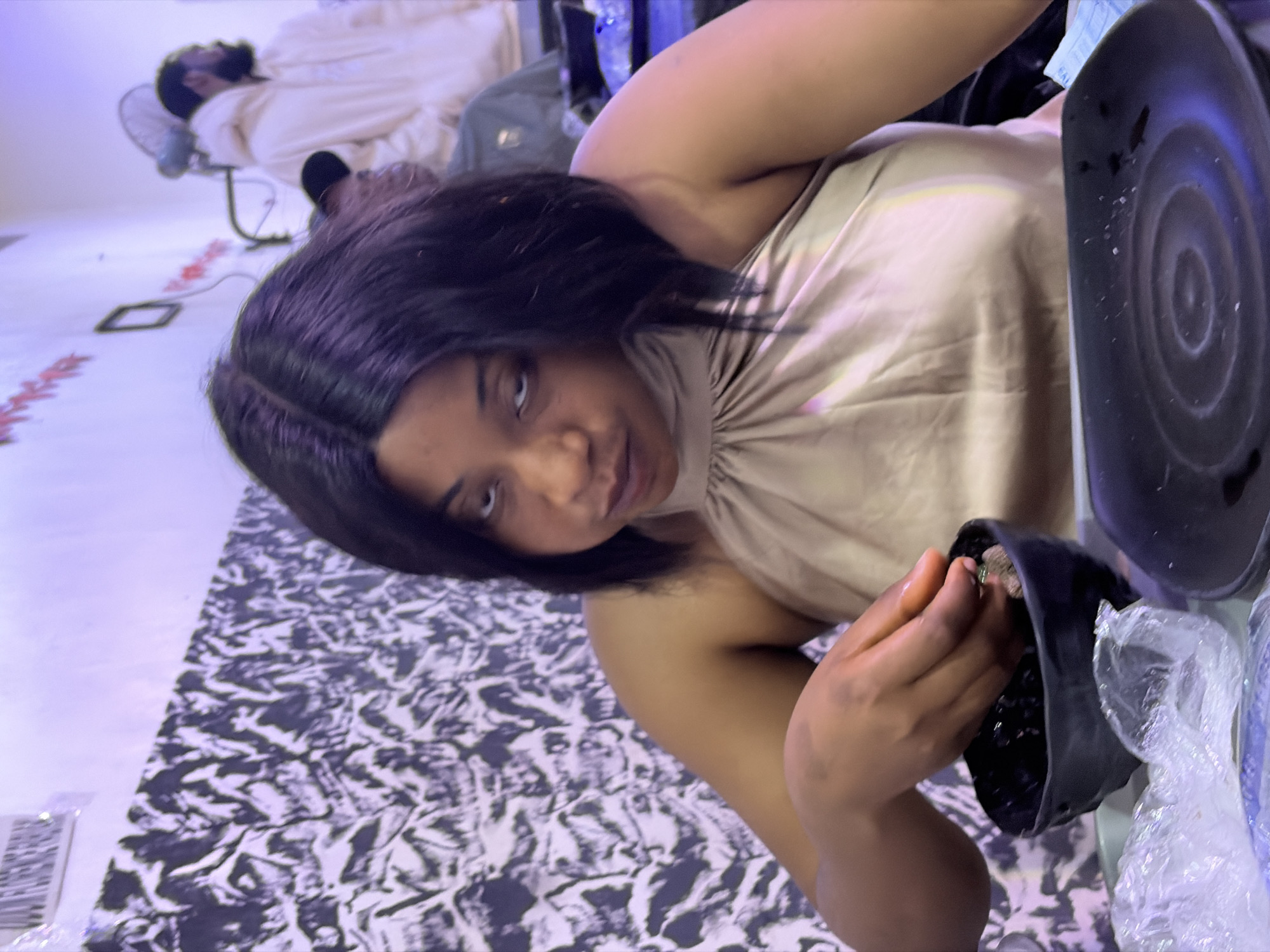 A medium shot captures a woman with dark, shoulder-length hair looking directly at the camera with a somewhat stern expression. She is wearing a sleeveless, light brown halter top that drapes around her neck. Her right hand is extended over a small, black bowl, and she appears to be picking something out of it with her fingers. The bowl contains dark material, possibly food. In front of her is a black plate with a circular pattern, and to her left, some plastic wrap is visible.
The background is dimly lit and features a wall with a black and white patterned wallpaper. To the right, another person in a light-colored hooded garment is visible, with a ceiling fan mounted above. There are some red decorations on the wall behind them. The overall lighting suggests an indoor setting, possibly a restaurant or eatery in Jos, Nigeria, indicated by the context provided. The atmosphere seems casual.