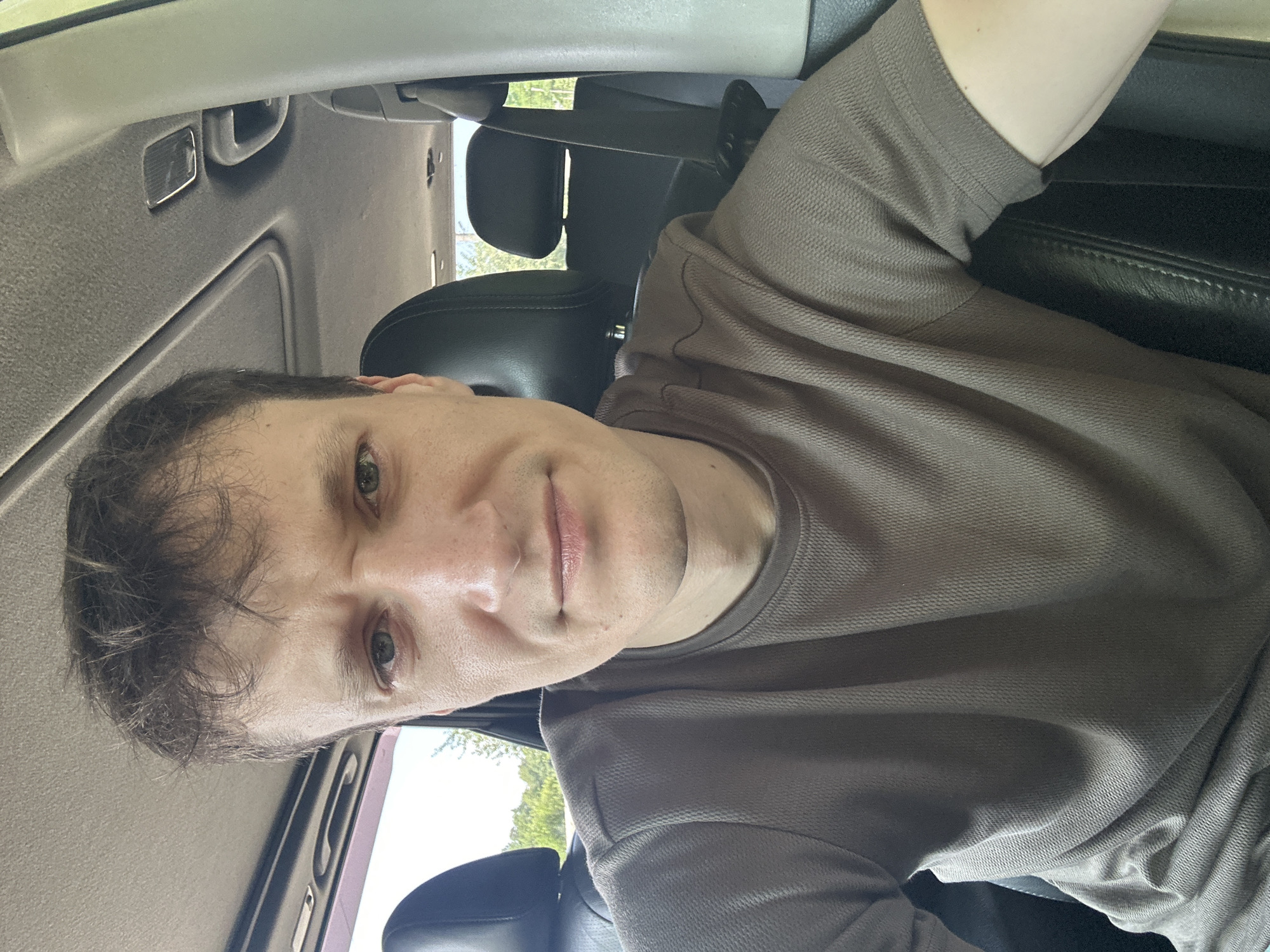 A selfie shows a man sitting in the driver's seat of a car. He is wearing a brown t-shirt. The car's interior is visible, including the headrests of the seats and the roof liner. Sunlight shines through the car windows, illuminating the scene. The image appears to have been taken outdoors, likely in a rural or suburban area, given the greenery visible through the windows. There is no specific indication of the location being in the city of Makiv, Ukraine, nor any context to suggest a crime or forensic scene.