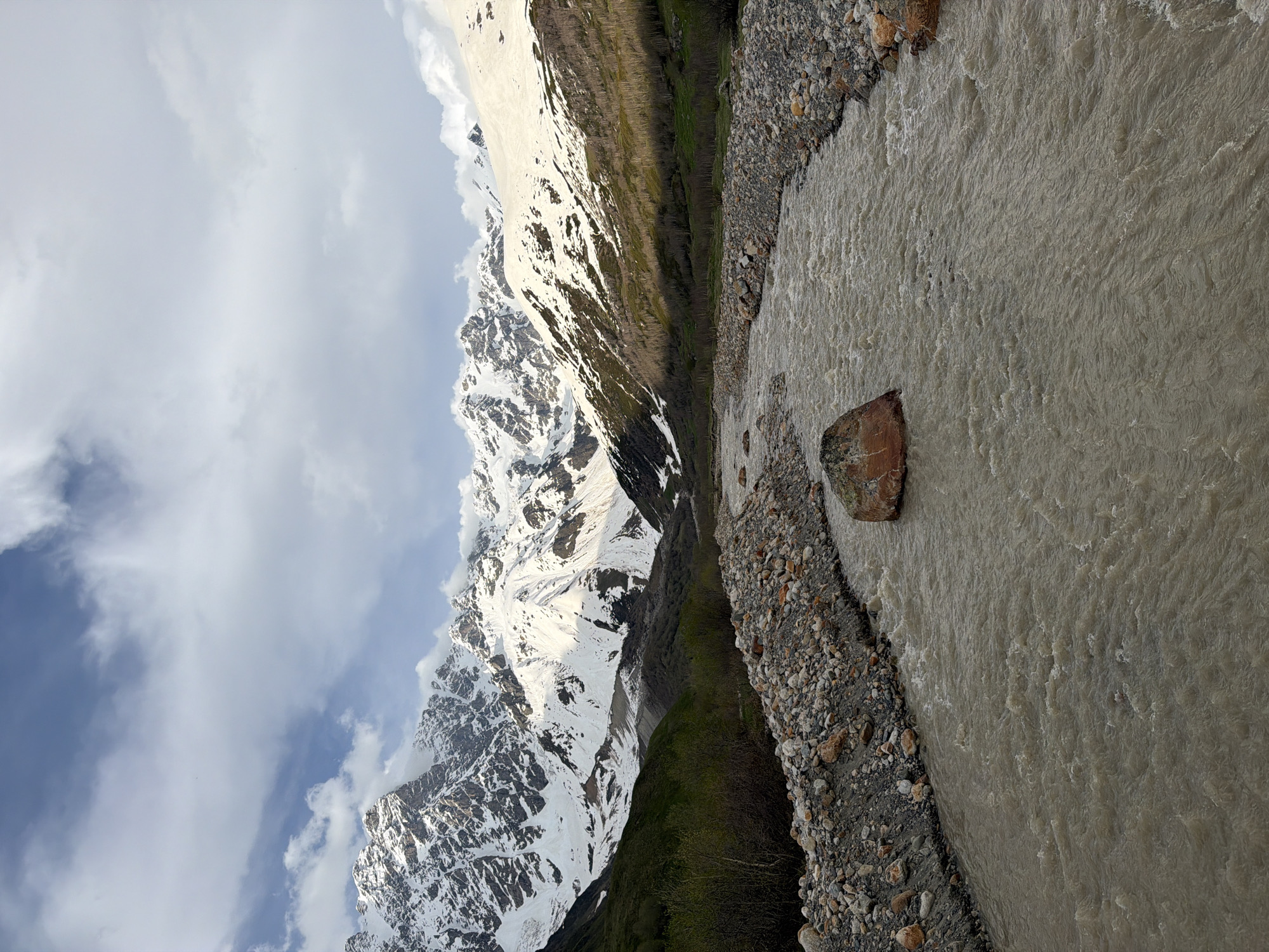 The image depicts a wide, muddy river flowing through a valley in Ushguli, Georgia. The riverbanks are lined with a mixture of small stones and larger rocks. In the foreground, a large, reddish-brown boulder sits in the middle of the river. The background is dominated by a range of snow-capped mountains, with some areas of green vegetation visible on the lower slopes. The sky is partly cloudy, with patches of blue visible between the white clouds. The overall impression is one of a rugged, natural landscape.