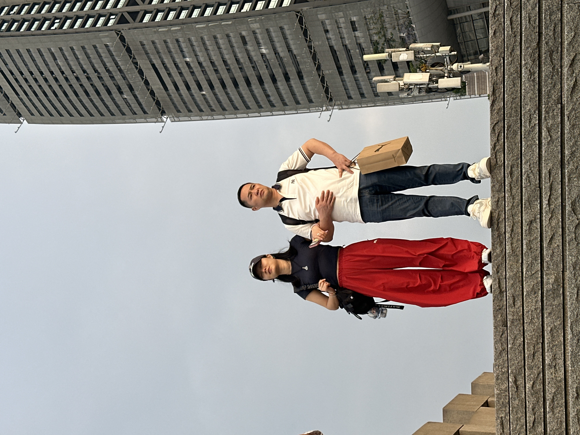 In Nan'an District, China, a man and a woman are standing outdoors against a neutral sky and the backdrop of a modern building. The man is wearing a white polo shirt with dark trim, dark jeans, and white sneakers, and he's holding a brown paper bag. The woman is dressed in a dark short-sleeved shirt, red pants, and appears to have a black bag slung over her shoulder. They are close to a stone wall and some steps.