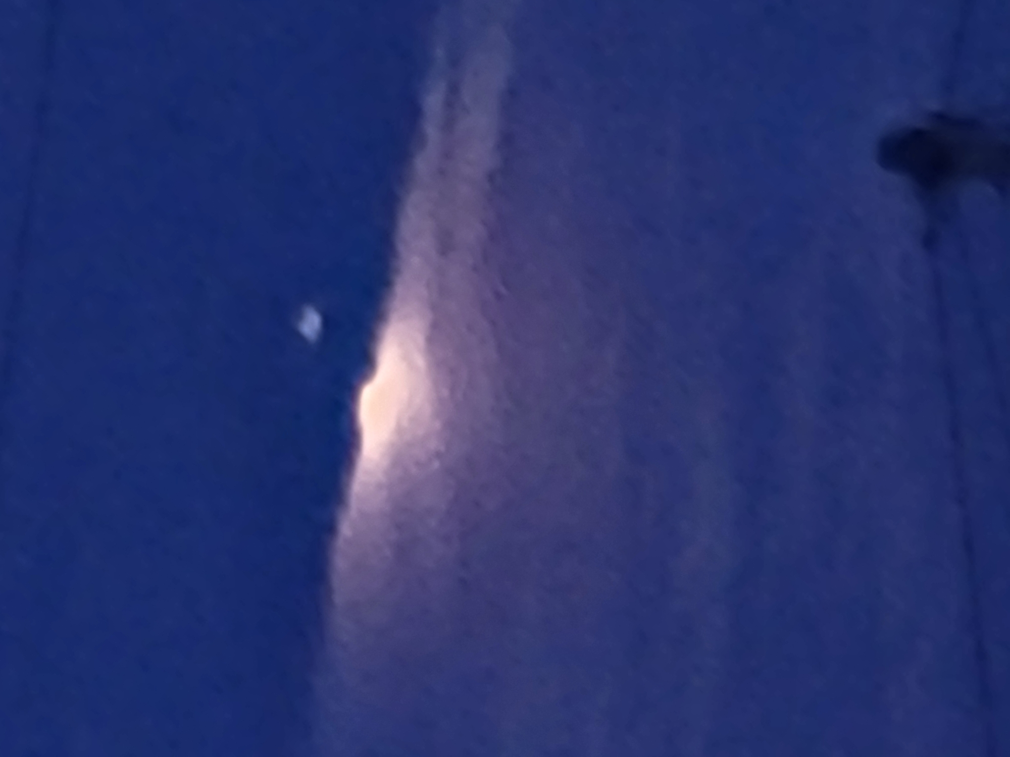 The image shows a blurry, dark blue sky with a bright, diffused light source in the upper middle. Below this light, there is a dark, horizontal band that appears to be clouds or a structure. Towards the bottom of the image, a faint, blurred figure can be seen, with outstretched arms, positioned on what appears to be a dark surface with some thin, horizontal lines. The overall scene is dark and lacks distinct details due to the blurriness and low light. There is no identifiable information to place the image in Jelgava, Latvia.
