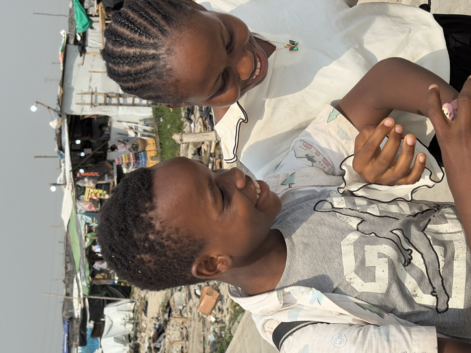 In Igboefon, Nigeria, a daylight scene captures two individuals, a boy and a young woman, in the foreground against a backdrop of an informal settlement. The boy, positioned on the left, has dark skin and short, dark, tightly curled hair. He wears a grey t-shirt featuring a prominent black outline of a jumping figure and the number "23," underneath a light-colored, short-sleeved button-up shirt with a subtle pattern. A black strap, likely from a backpack, is visible over his left shoulder. He is smiling, looking down intently at a small, pink object held in his left hand. The young woman, on the right, also has dark skin and dark hair styled in braids. She wears a white, short-sleeved top with a ruffled collar and trim, and a gold chain with a multi-colored butterfly pendant around her neck. She is smiling and looking down in the same direction as the boy, with her right hand positioned near his left arm.
The background reveals a dense collection of makeshift structures, indicative of an informal settlement. These buildings are constructed from various materials including corrugated iron, tarpaulins, and possibly wooden planks, suggesting a low-income residential or commercial area. Some structures appear to function as small shops or stalls, with bottled beverages and other goods visible. Debris and scattered materials are present on the ground. Overhead, utility poles with visible light fixtures extend above the structures, indicating some infrastructure. The sky is a uniform light grey, suggesting either overcast conditions or a hazy, bright day.