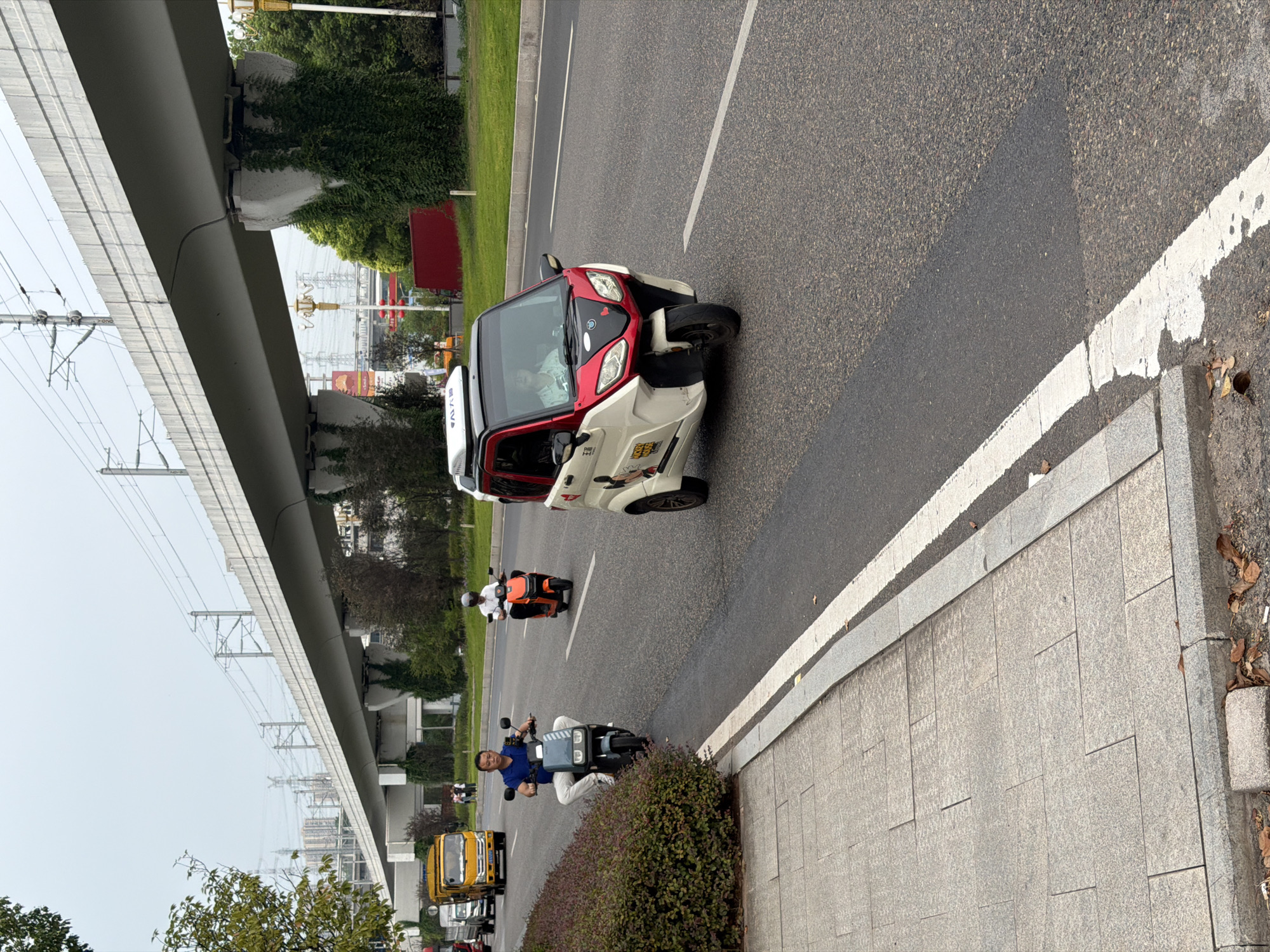 The image depicts a street scene in Jiangjin District, Chongqing, China, under an elevated railway or highway. In the foreground, a sidewalk made of granite slabs runs parallel to a street. On the street, a man in a blue shirt and white pants rides a grey electric scooter. Further down the road, an orange electric scooter is ridden by a person wearing a white helmet and white shirt. A white and red three-wheeled microcar with Mickey Mouse decals is also visible on the road. The background shows buildings, trees, and overhead electrical wires associated with the elevated structure. The sky is overcast, indicating a cloudy day. The overall environment suggests an urban setting with active transportation.